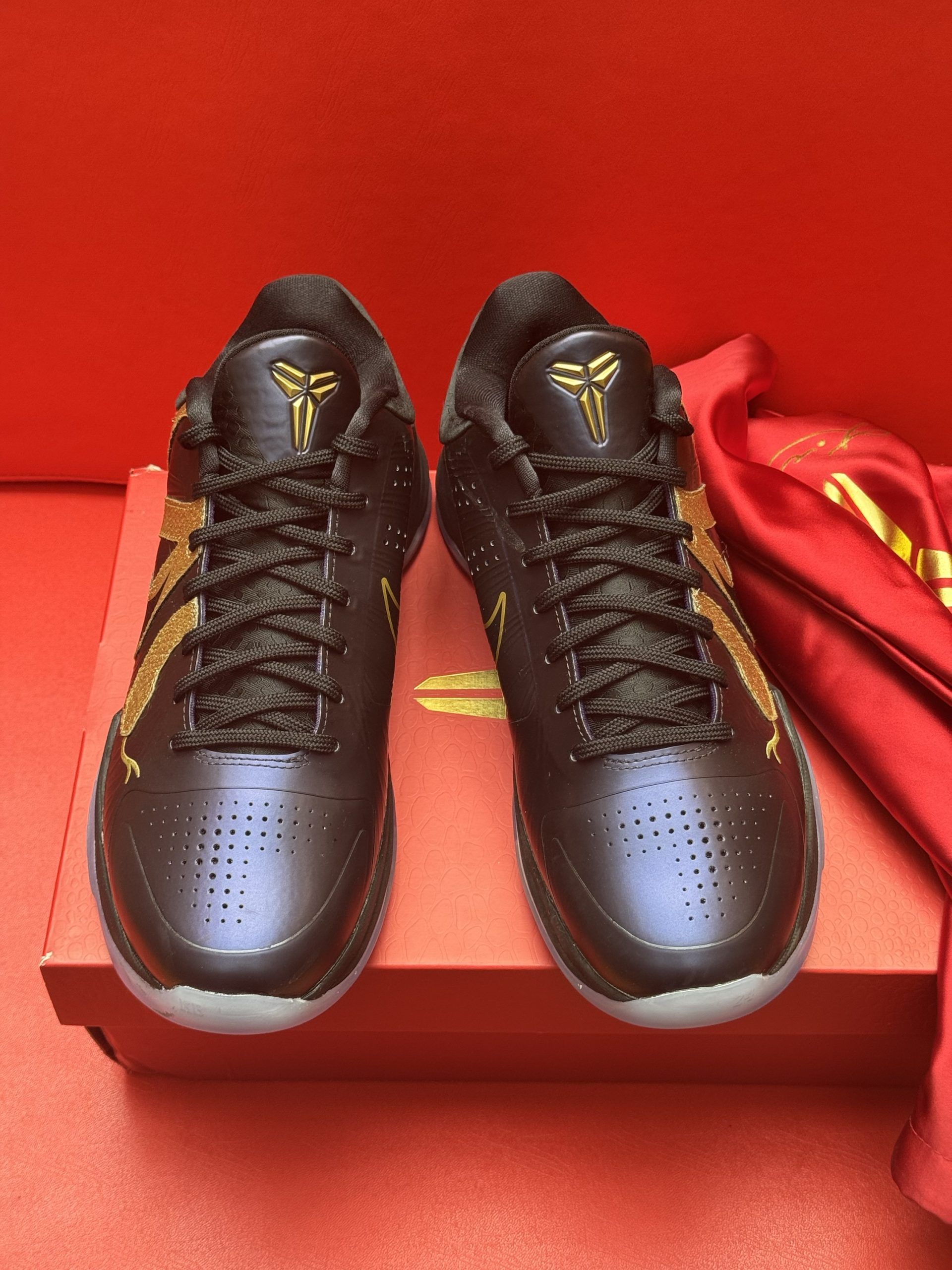 Black and gold Nike Kobe sneakers with a snake design, on a red box with a red cloth, a red background.
