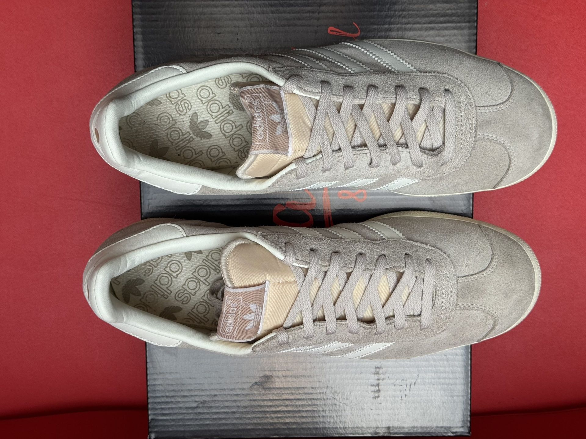 Light gray Adidas Gazelle sneakers with white stripes, on top of a black box, with the sole visible.