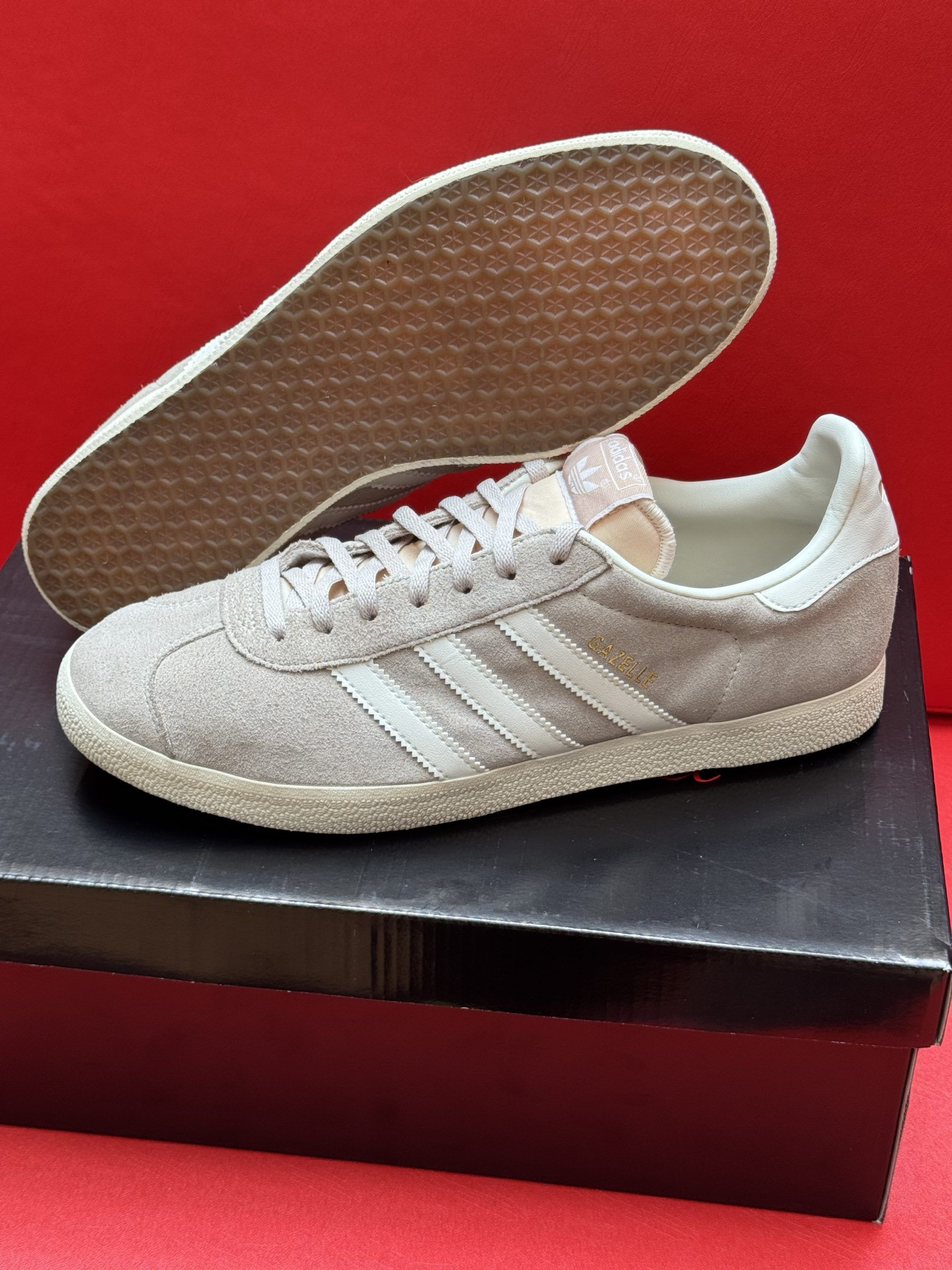 Light gray Adidas Gazelle sneakers with white stripes, on top of a black box, with the sole visible.
