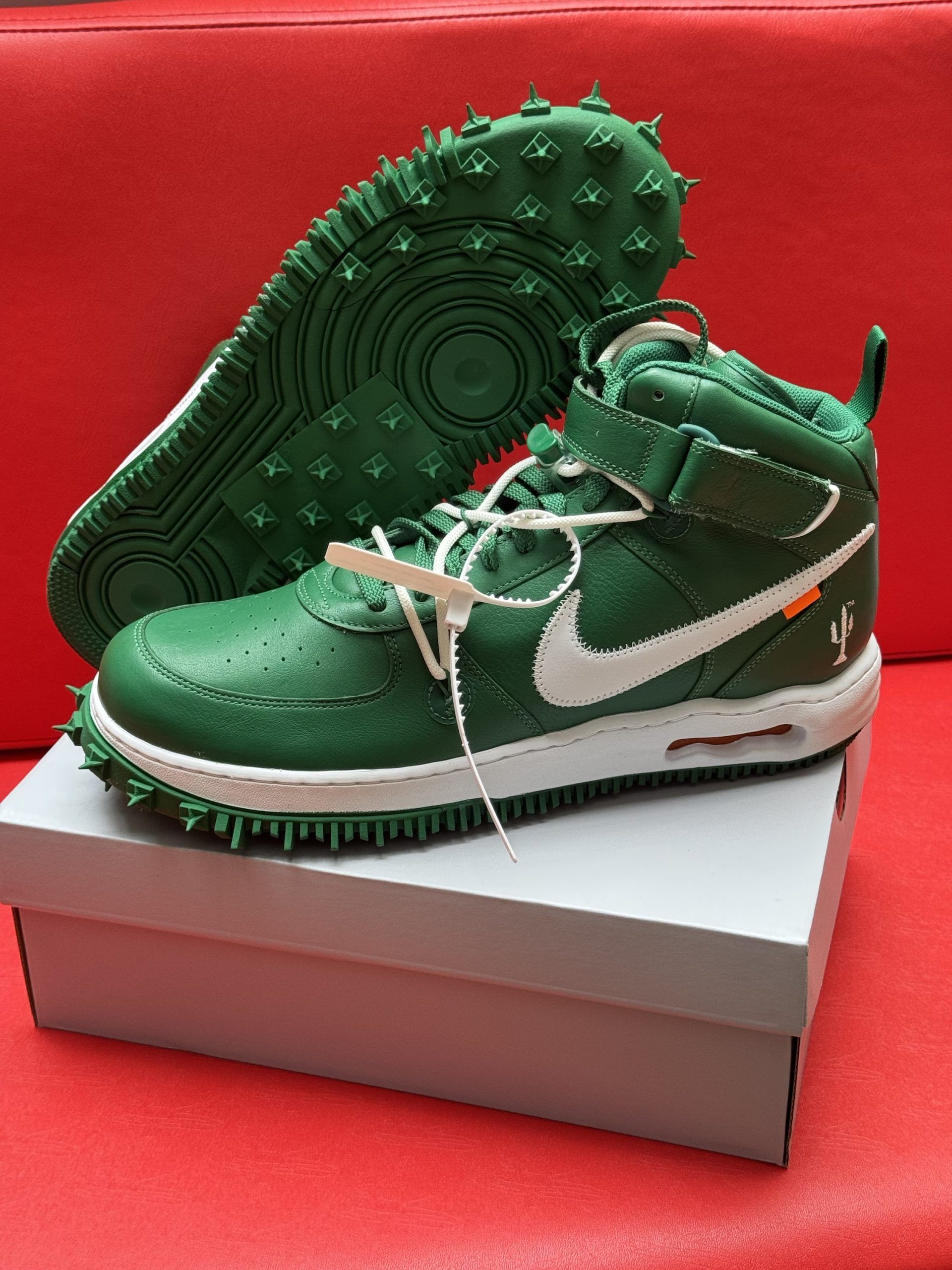 Green and white Off-White Nike Air Force 1 sneakers with spiked soles, on a white box against a red background.