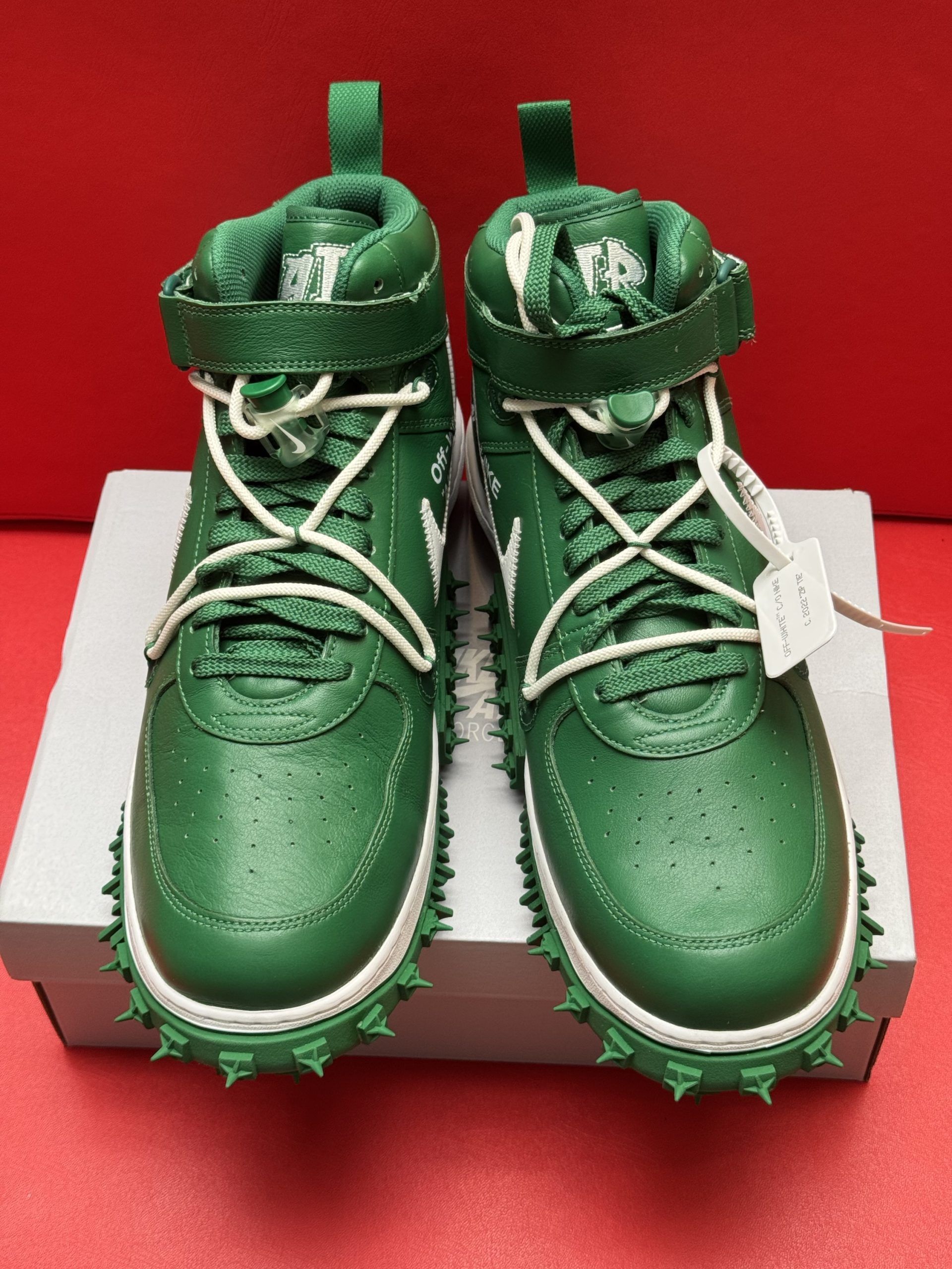 Green and white Off-White Nike Air Force 1 sneakers with spiked soles, on a white box against a red background.