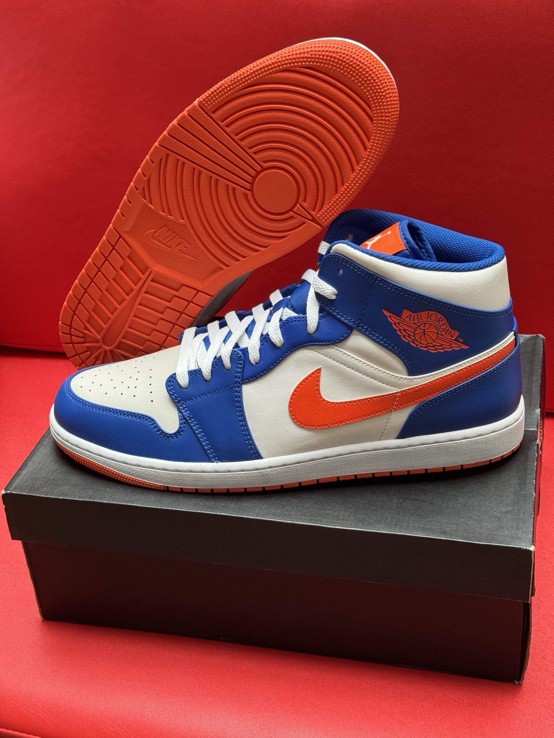 Blue, white, and orange Air Jordan 1 Mid sneaker on a black shoebox, with matching orange sole in the background.
