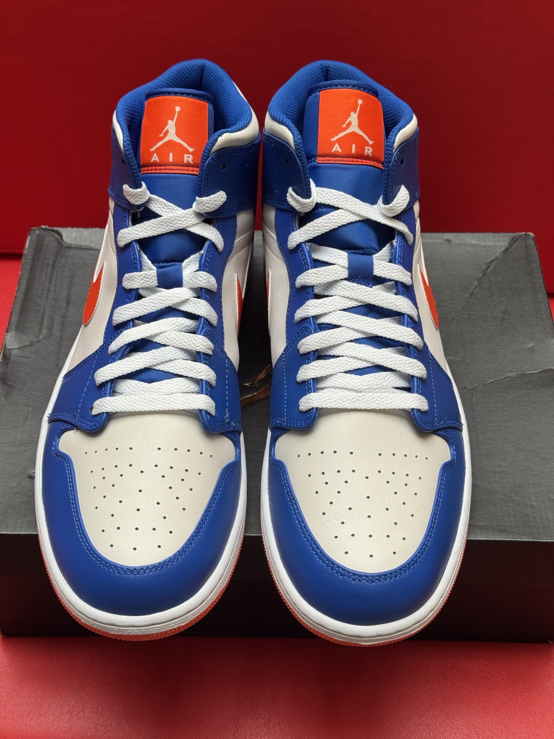 Blue, white, and orange Air Jordan 1 Mid sneaker on a black shoebox, with matching orange sole in the background.