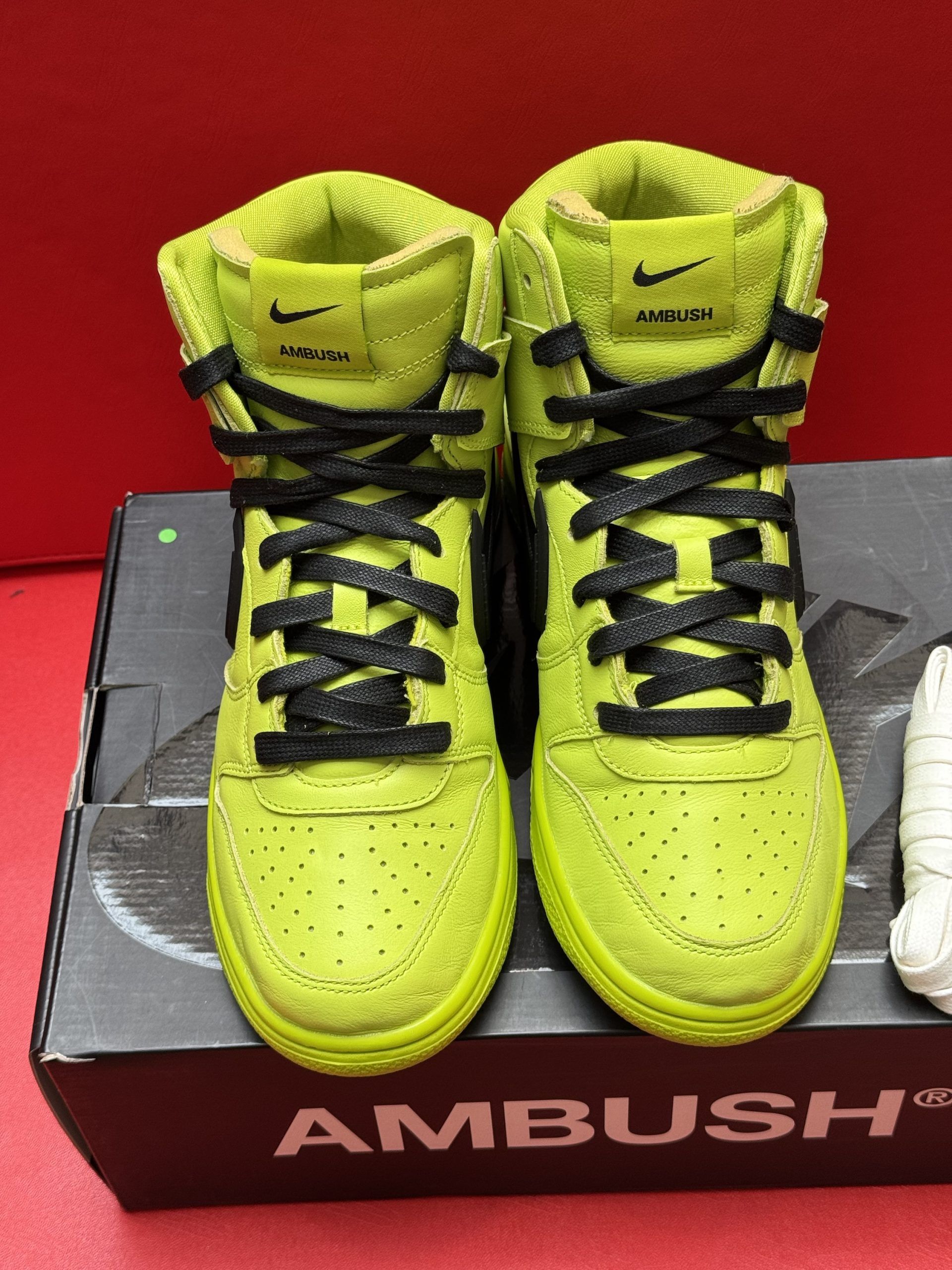 Green Nike high-top sneakers with black laces, a black swoosh, and a neon green sole, sitting on an AMBUSH shoe box.