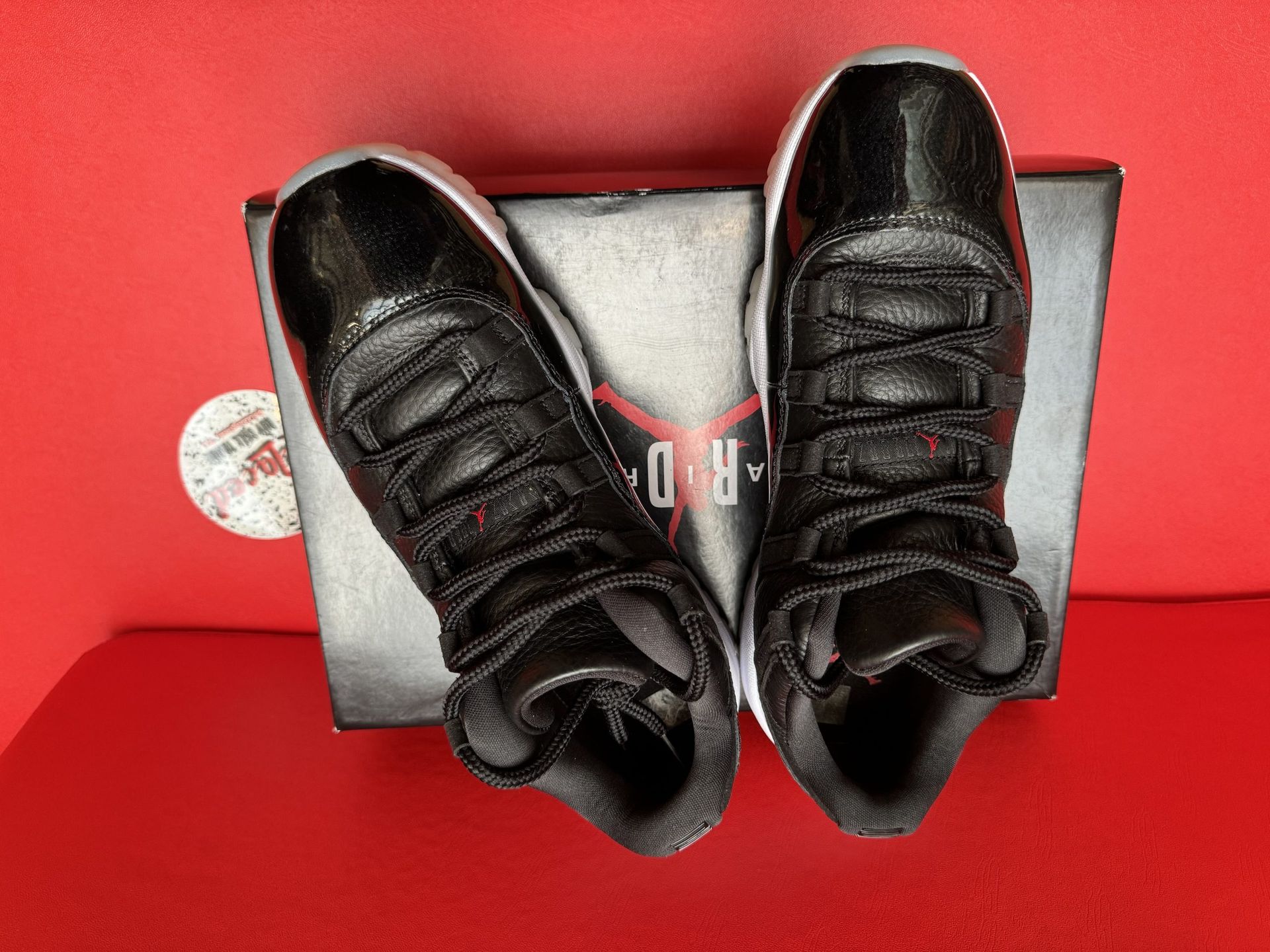 Black and white toddler sneakers with shiny material, white sole, and black laces, on a black box, against a red background.