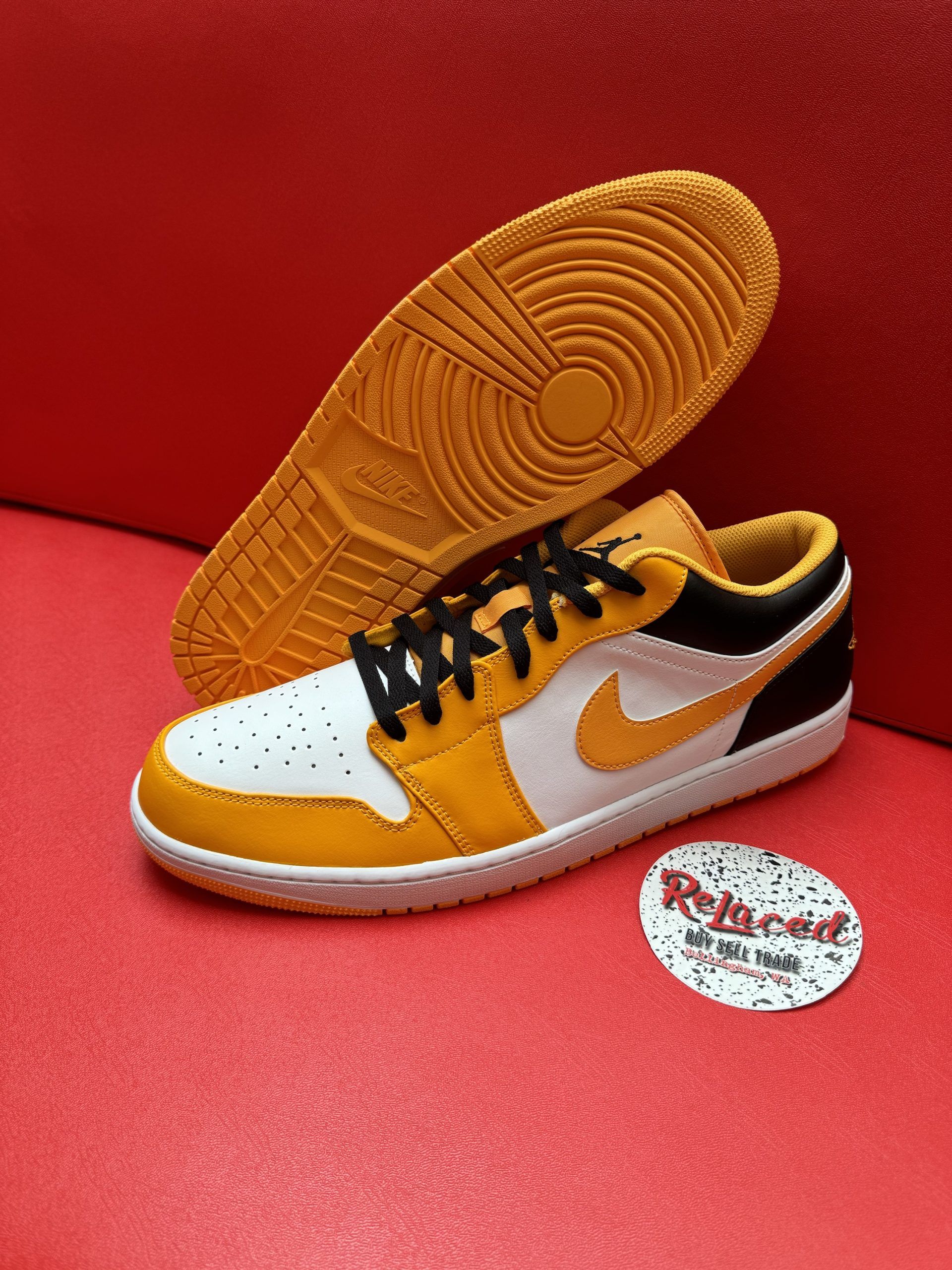 Yellow and white Air Jordan 1 Low sneaker with black laces, sole up, on a red surface.