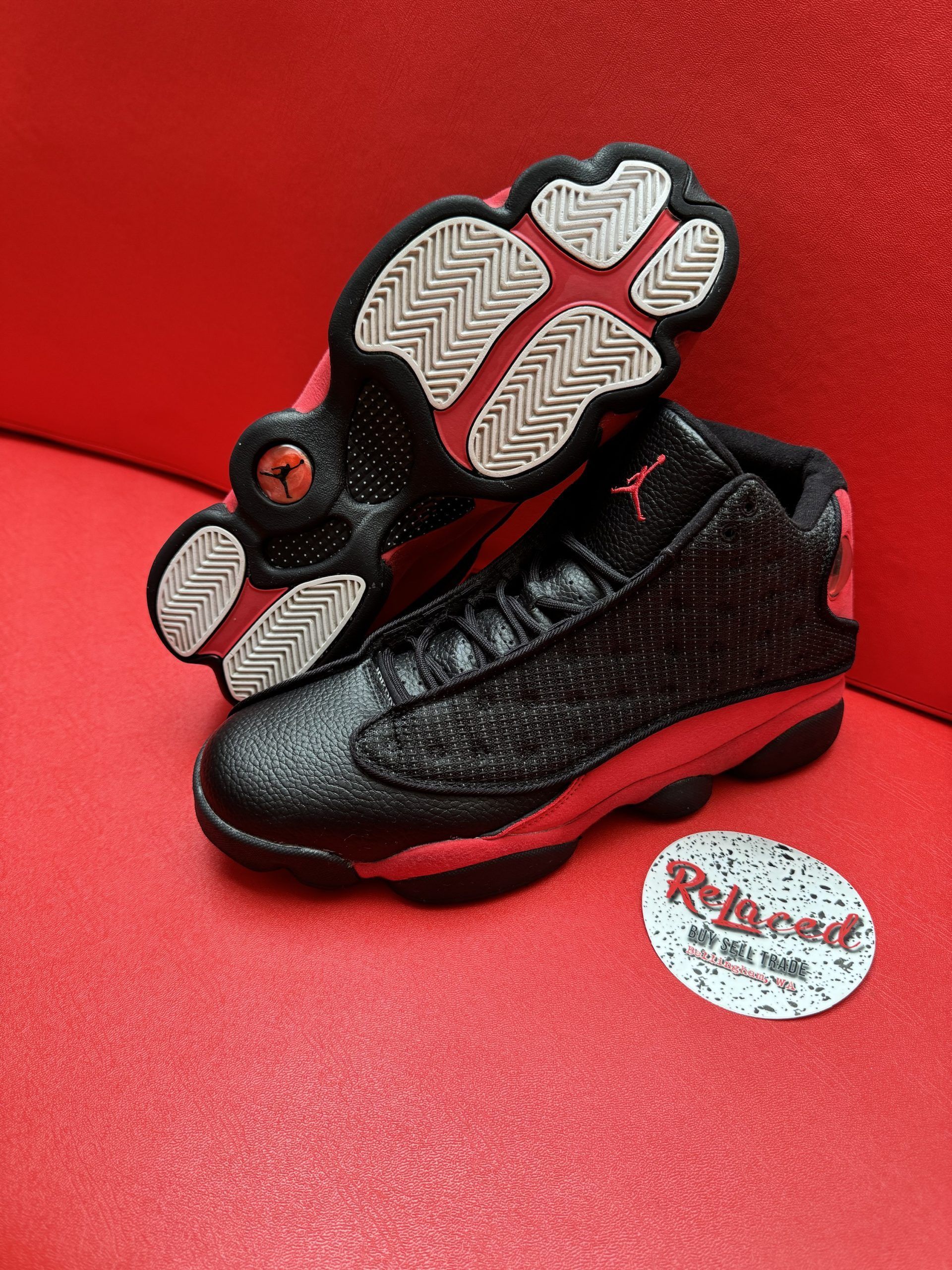 Black and red Air Jordan 13 sneakers on a red surface, with a logo sticker.
