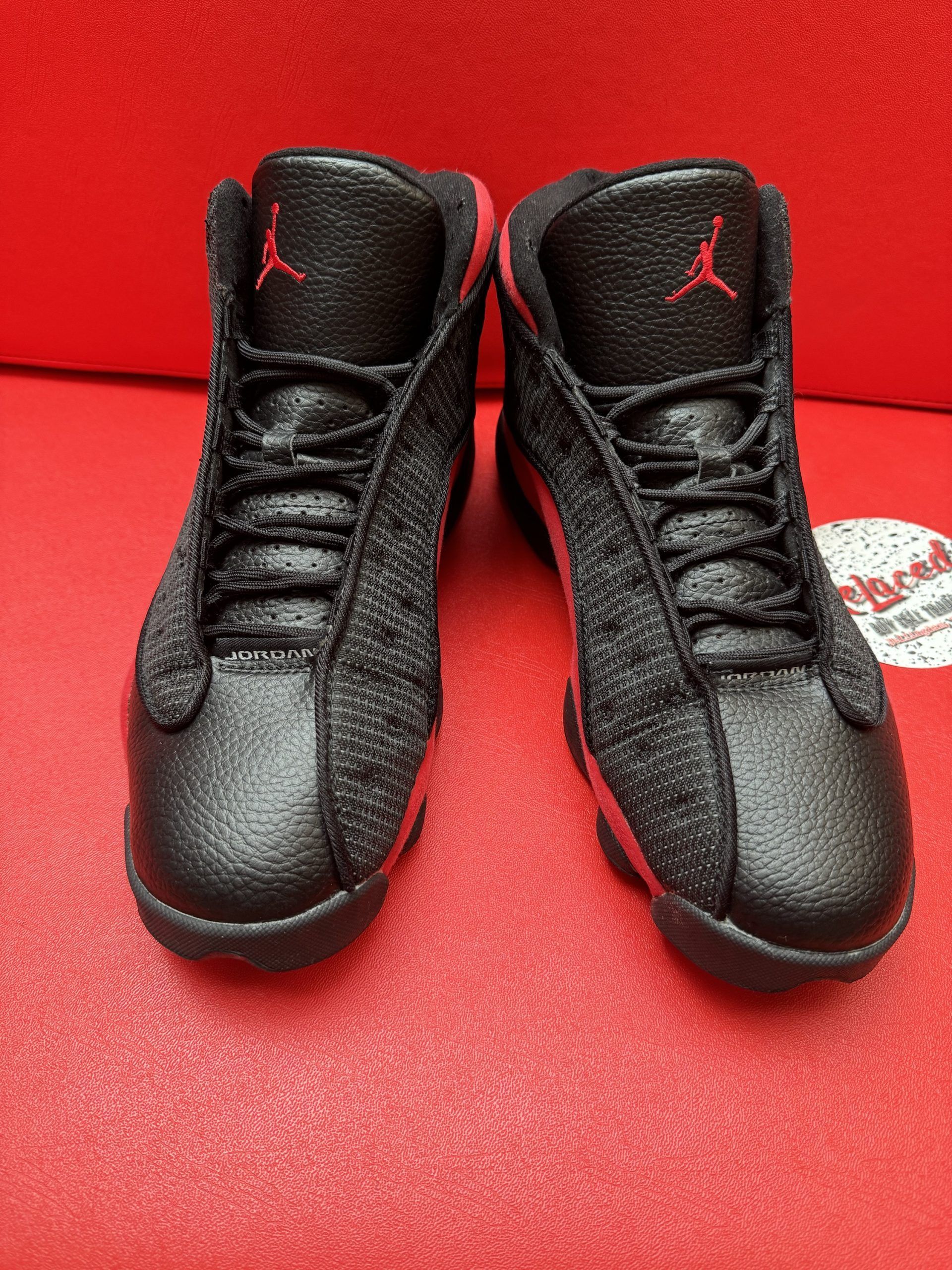 Black and red Air Jordan 13 sneakers on a red surface, with a logo sticker.