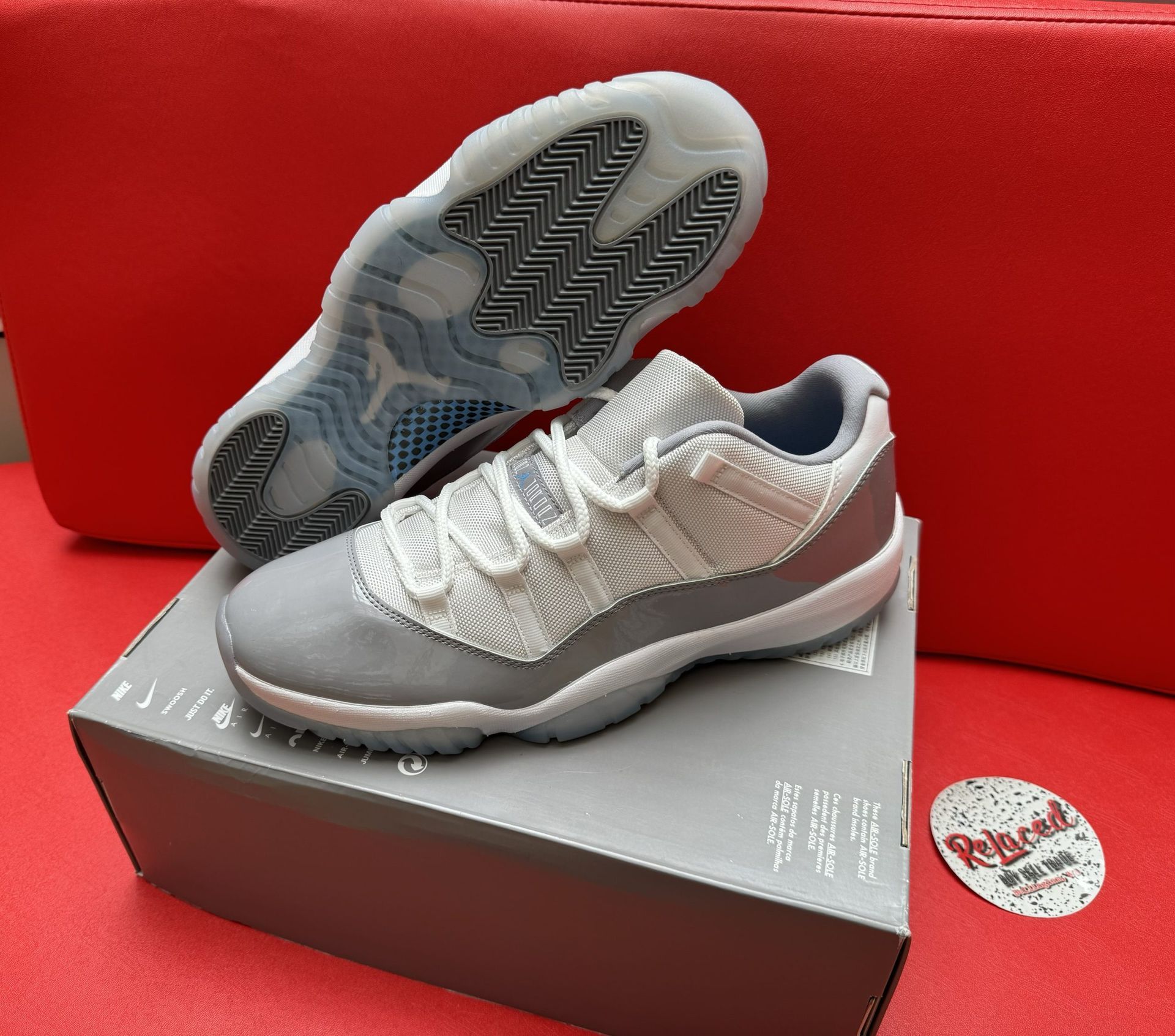 Gray and white Air Jordan 11 sneakers on a box, with a clear sole, against a red backdrop.