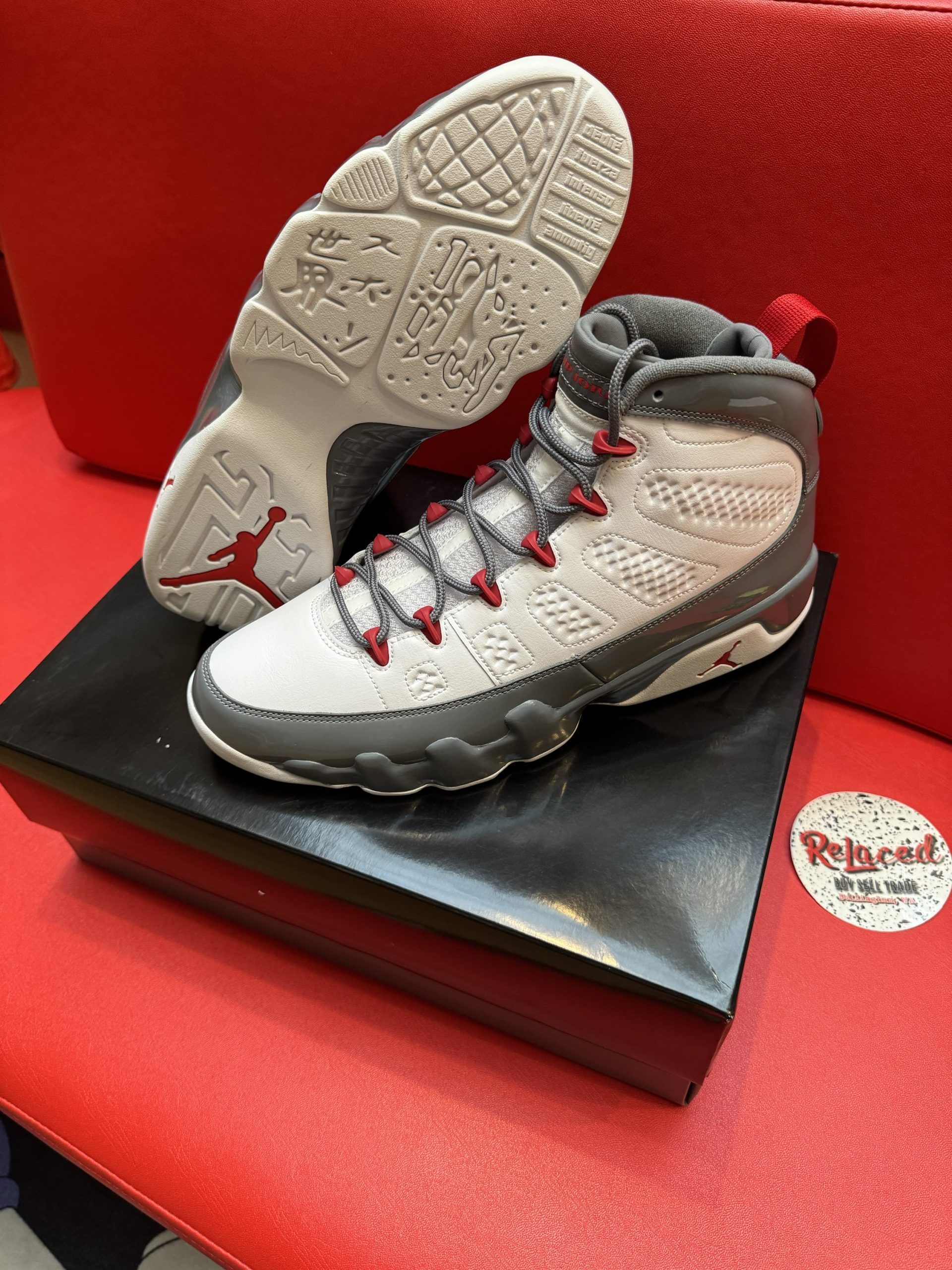 Air Jordan 9 sneakers, white and gray with red accents, on a black box. The sole of one shoe is visible.