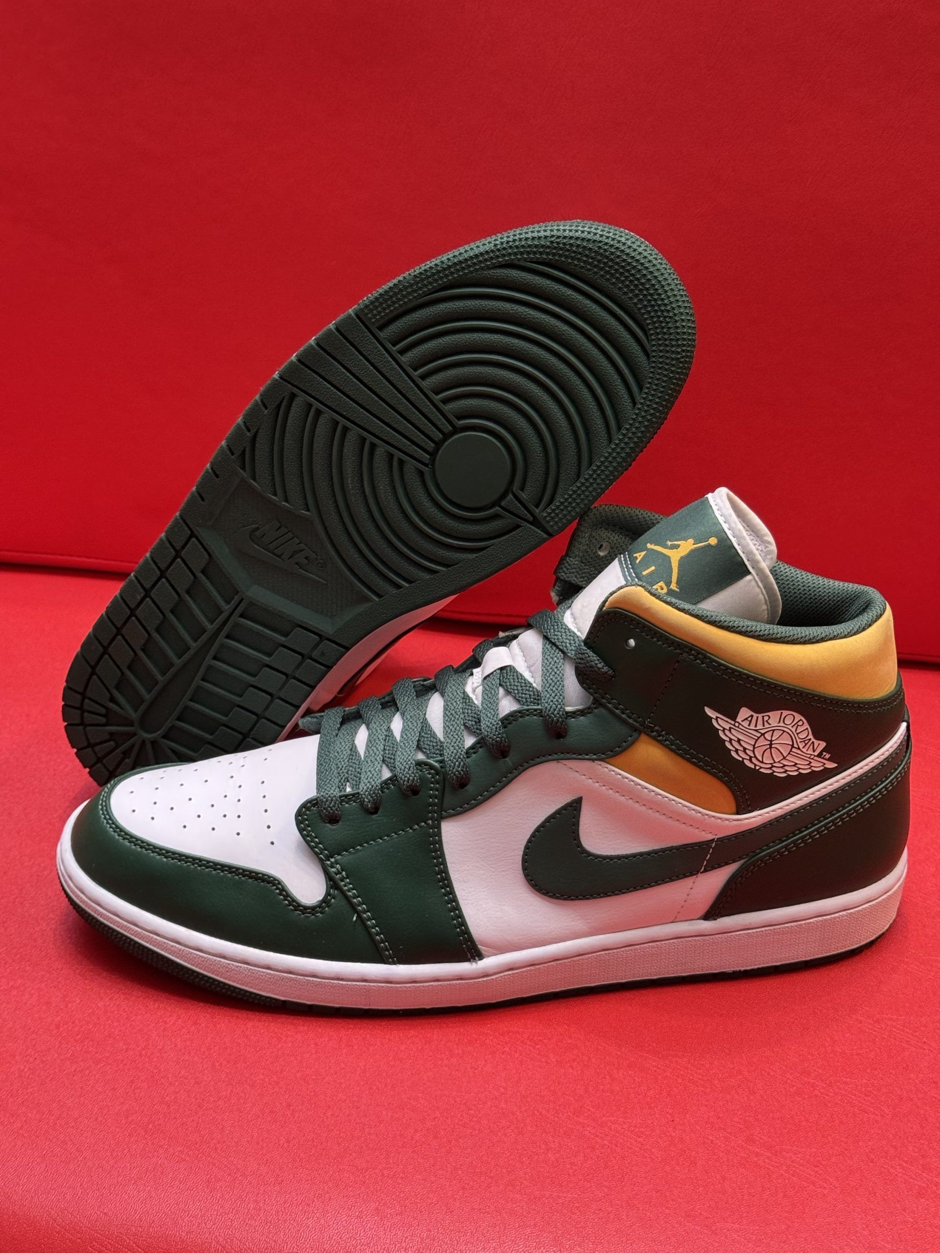 Green, white, and yellow Air Jordan 1 Mid sneaker on red surface. Sole visible behind.