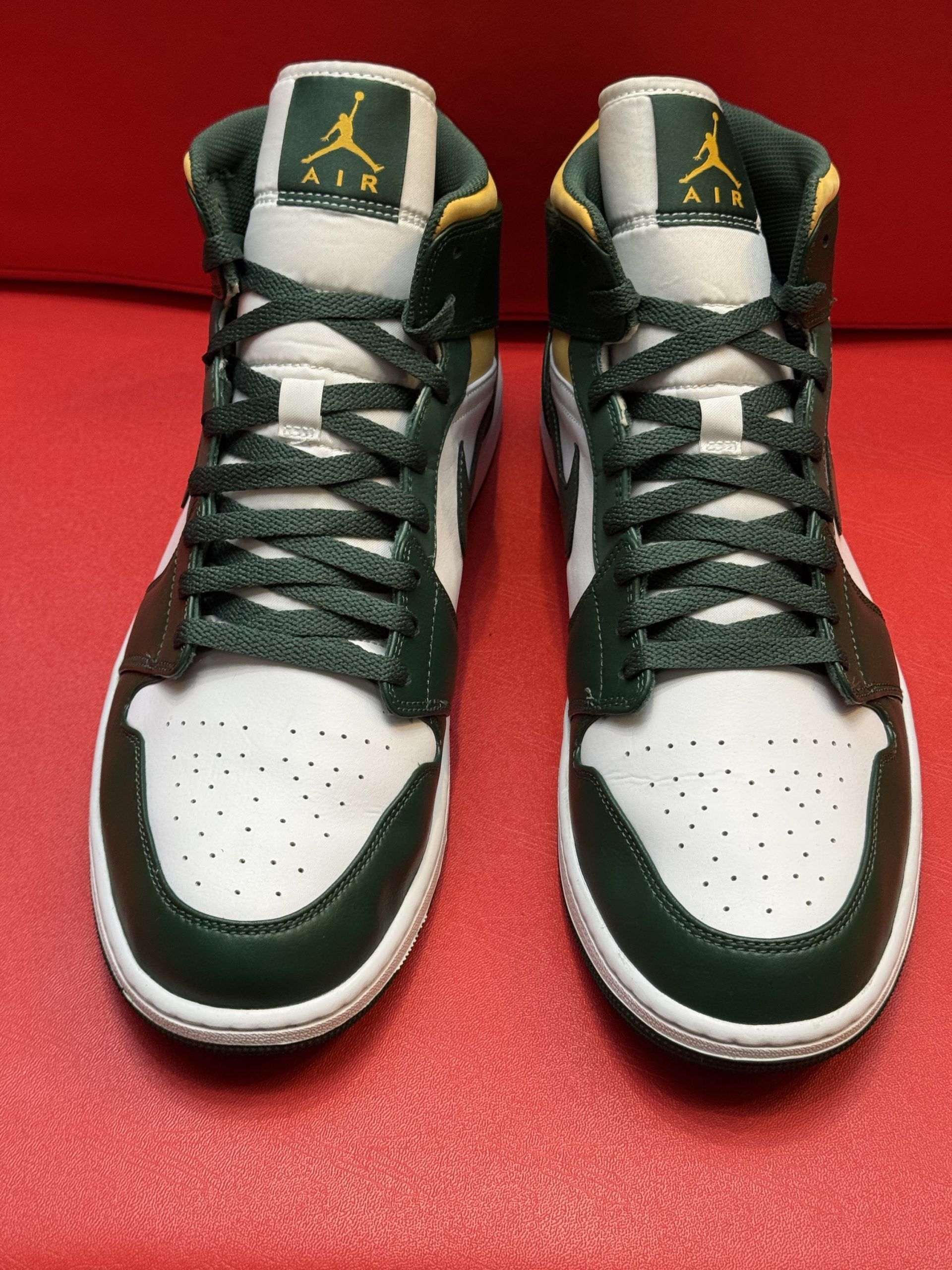 Green, white, and yellow Air Jordan 1 Mid sneaker on red surface. Sole visible behind.