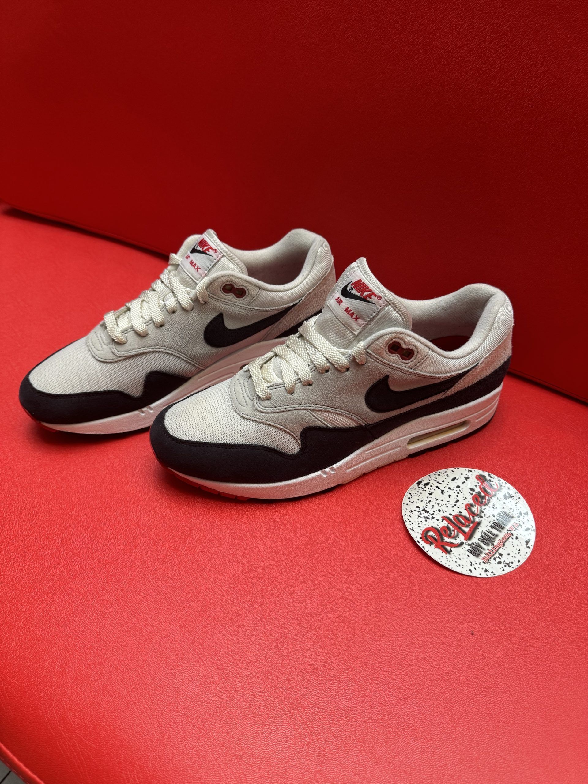 Pair of Nike Air Max 1 sneakers in gray, black, and red on a red surface.