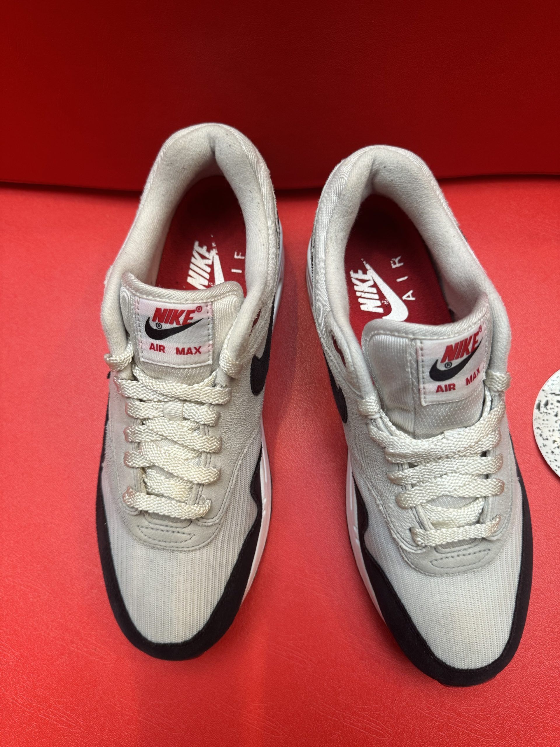 Pair of Nike Air Max 1 sneakers in gray, black, and red on a red surface.