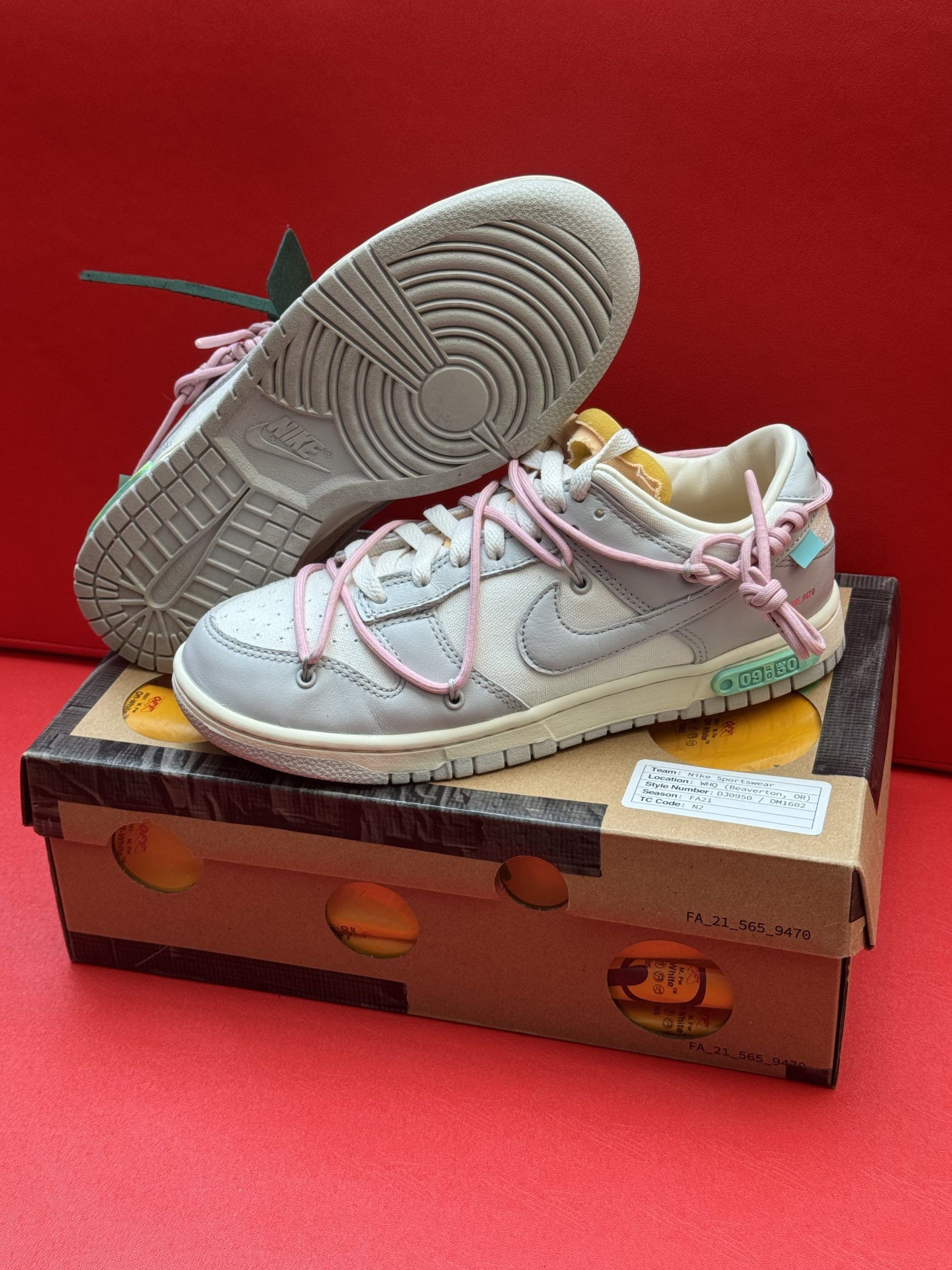 Off-White x Nike Dunk Low sneakers, grey with pink laces, on the shoe box. One shoe upside down. Red background.