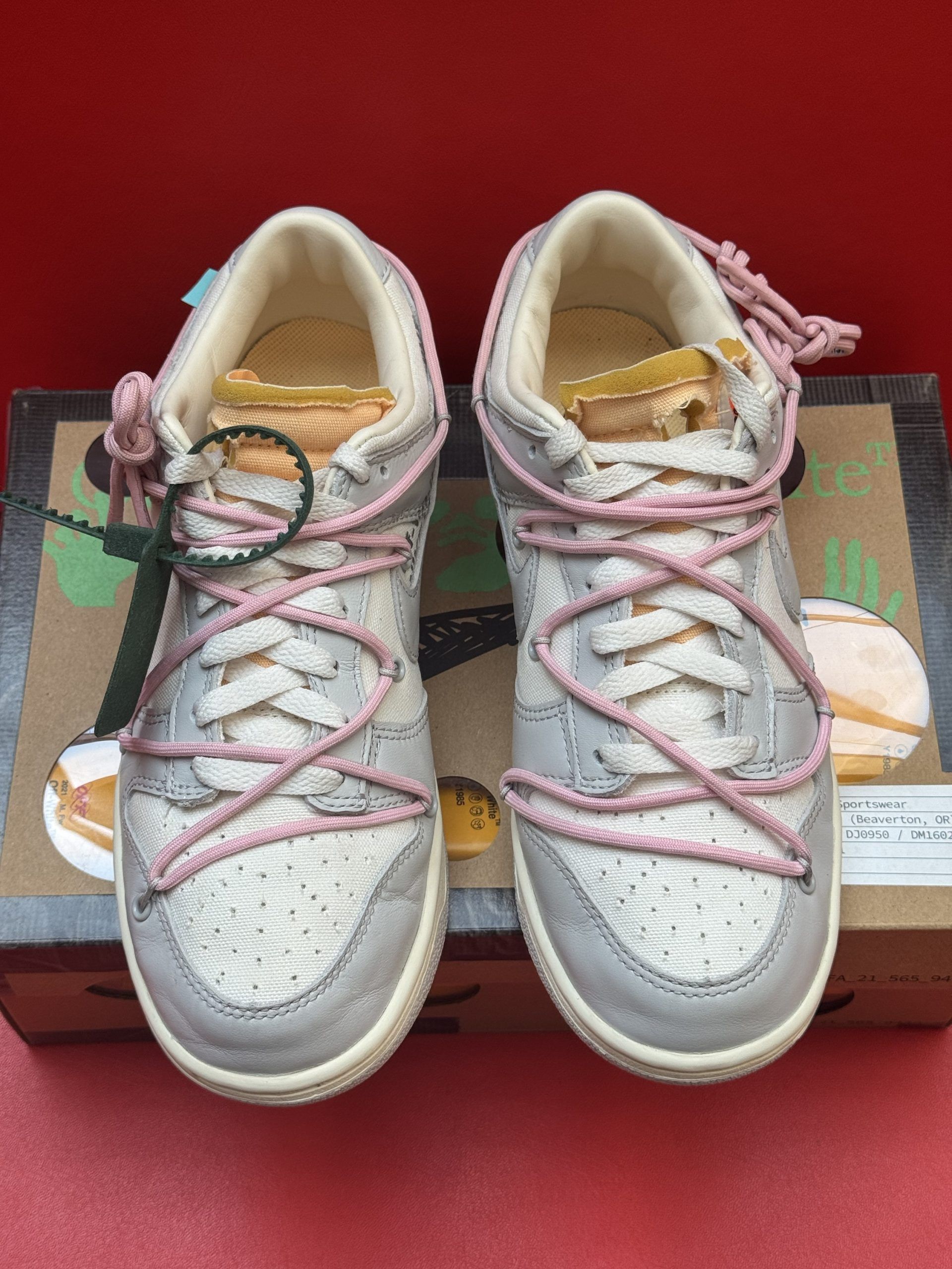 Off-White x Nike Dunk Low sneakers, grey with pink laces, on the shoe box. One shoe upside down. Red background.