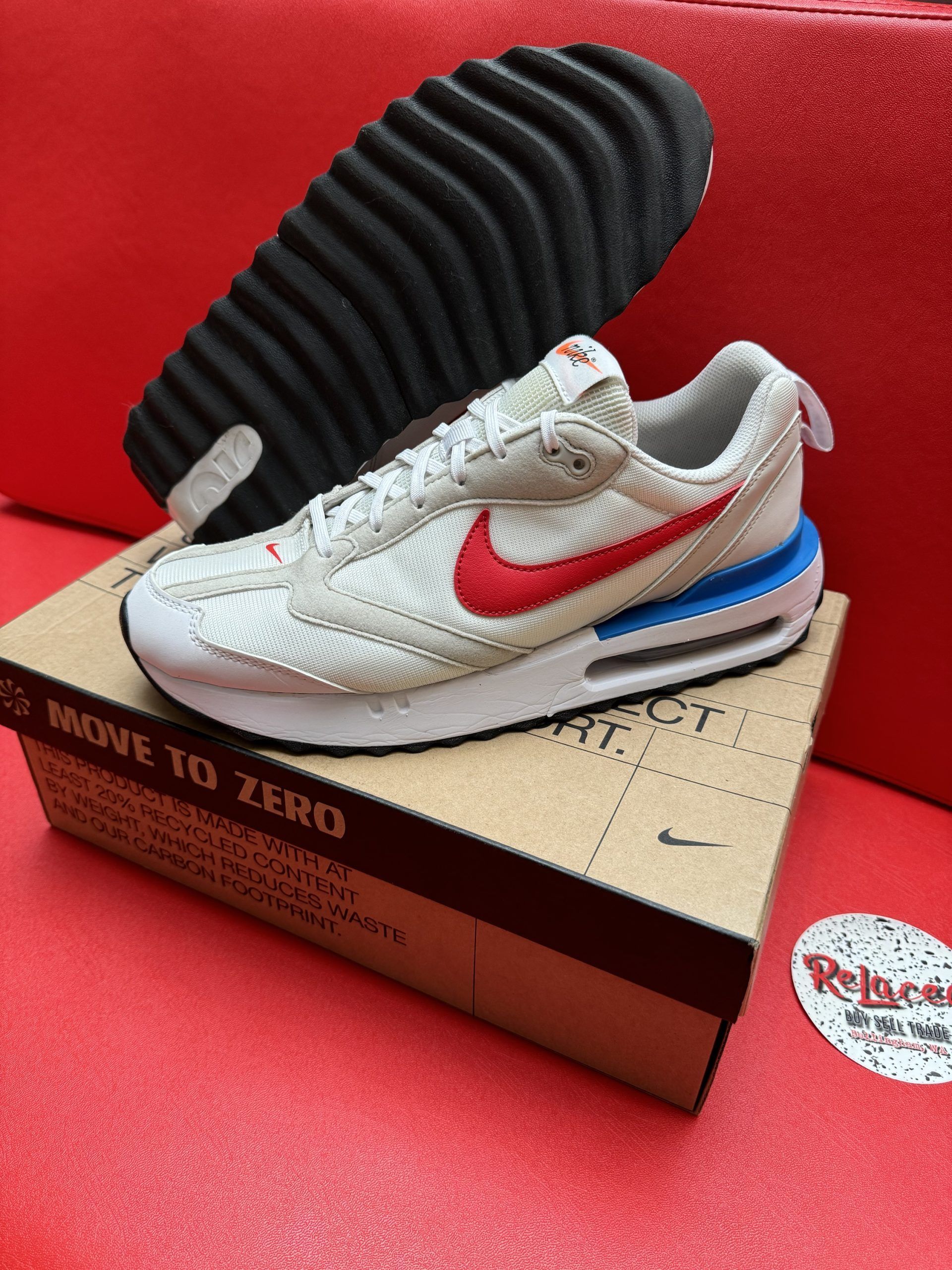 Beige Nike sneaker with red swoosh and blue midsole, box, and black outsole on red surface.