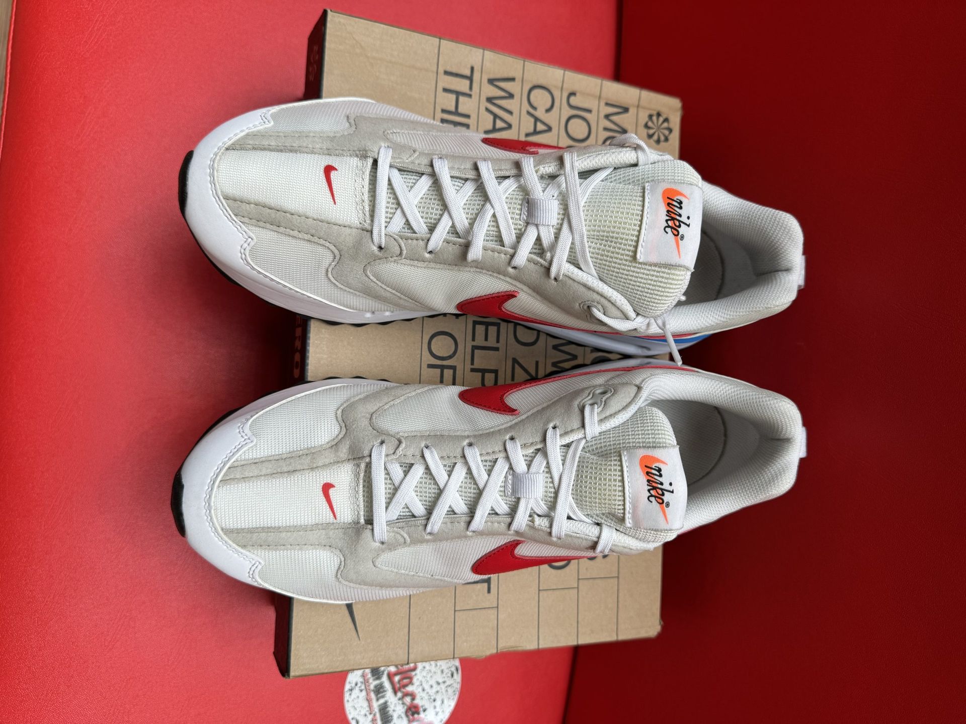 Beige Nike sneaker with red swoosh and blue midsole, box, and black outsole on red surface.