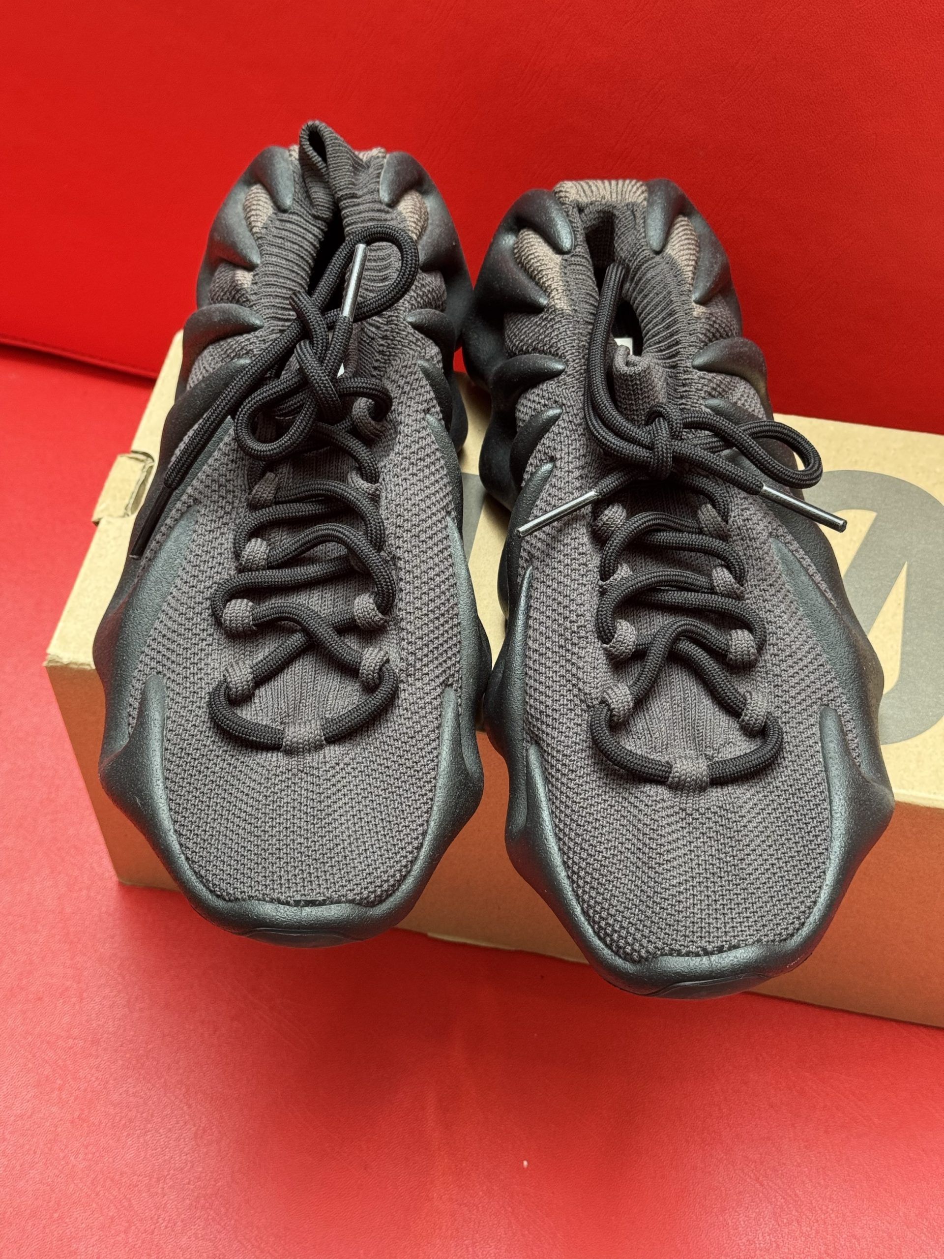 Yeezy 450 sneakers, dark gray with brown accents, on a brown cardboard box, set against a red background.