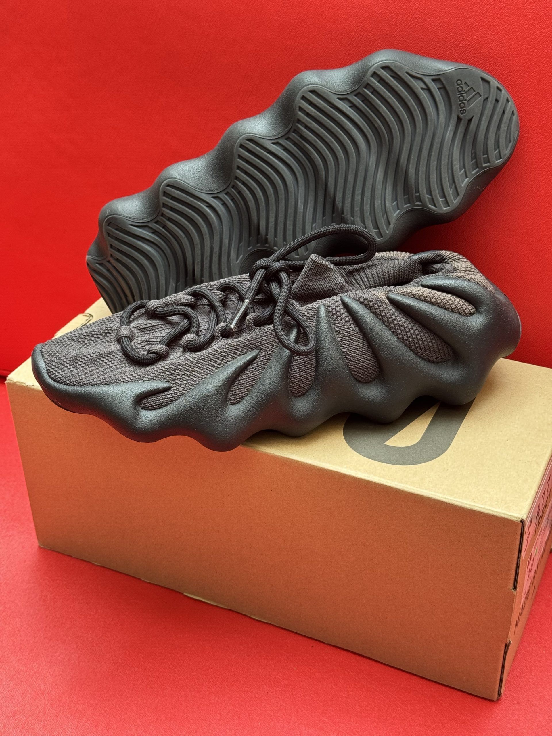 Yeezy 450 sneakers, dark gray with brown accents, on a brown cardboard box, set against a red background.