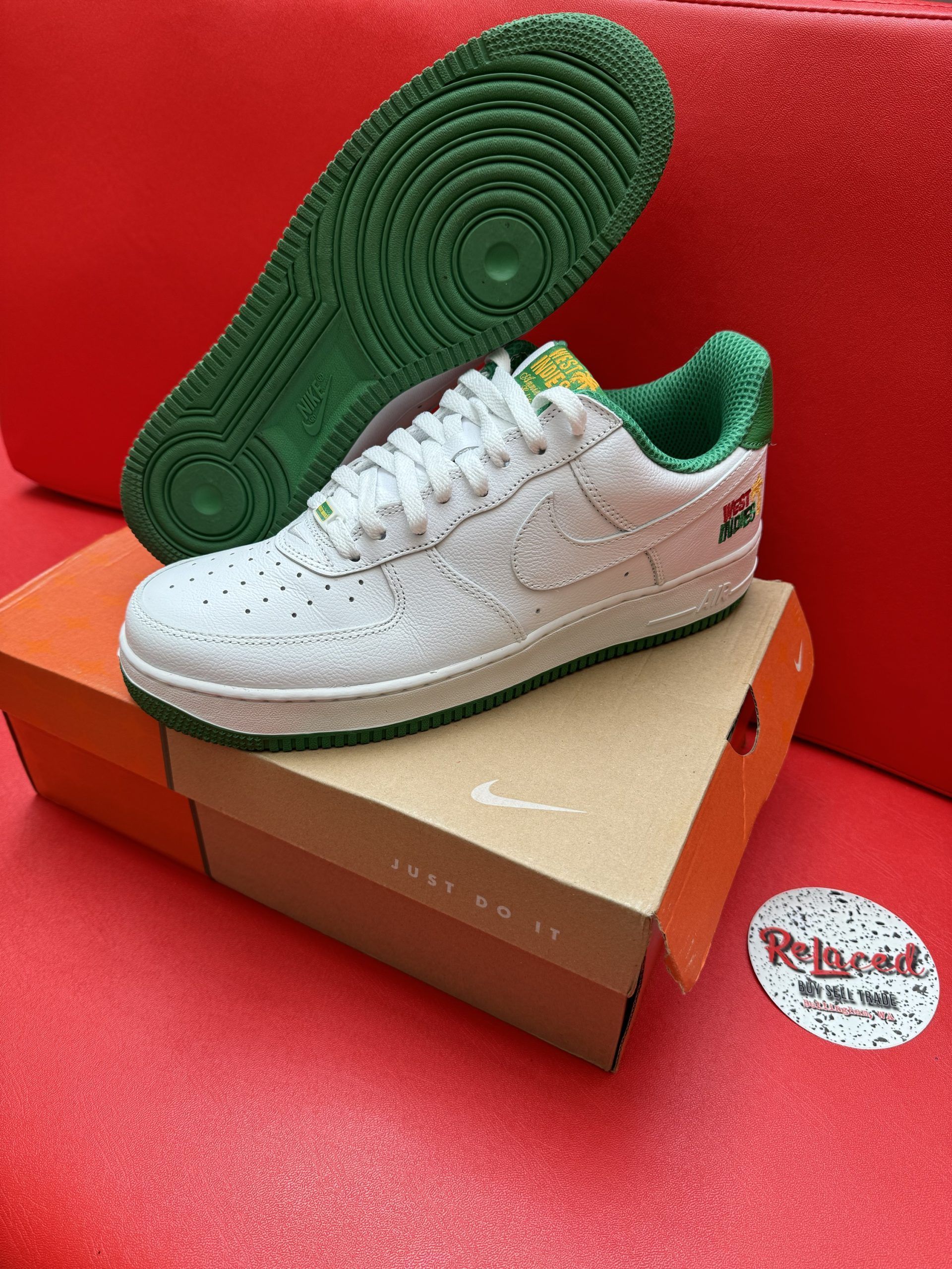 White Nike Air Force 1 sneaker with green accents, box, and sole on red surface.