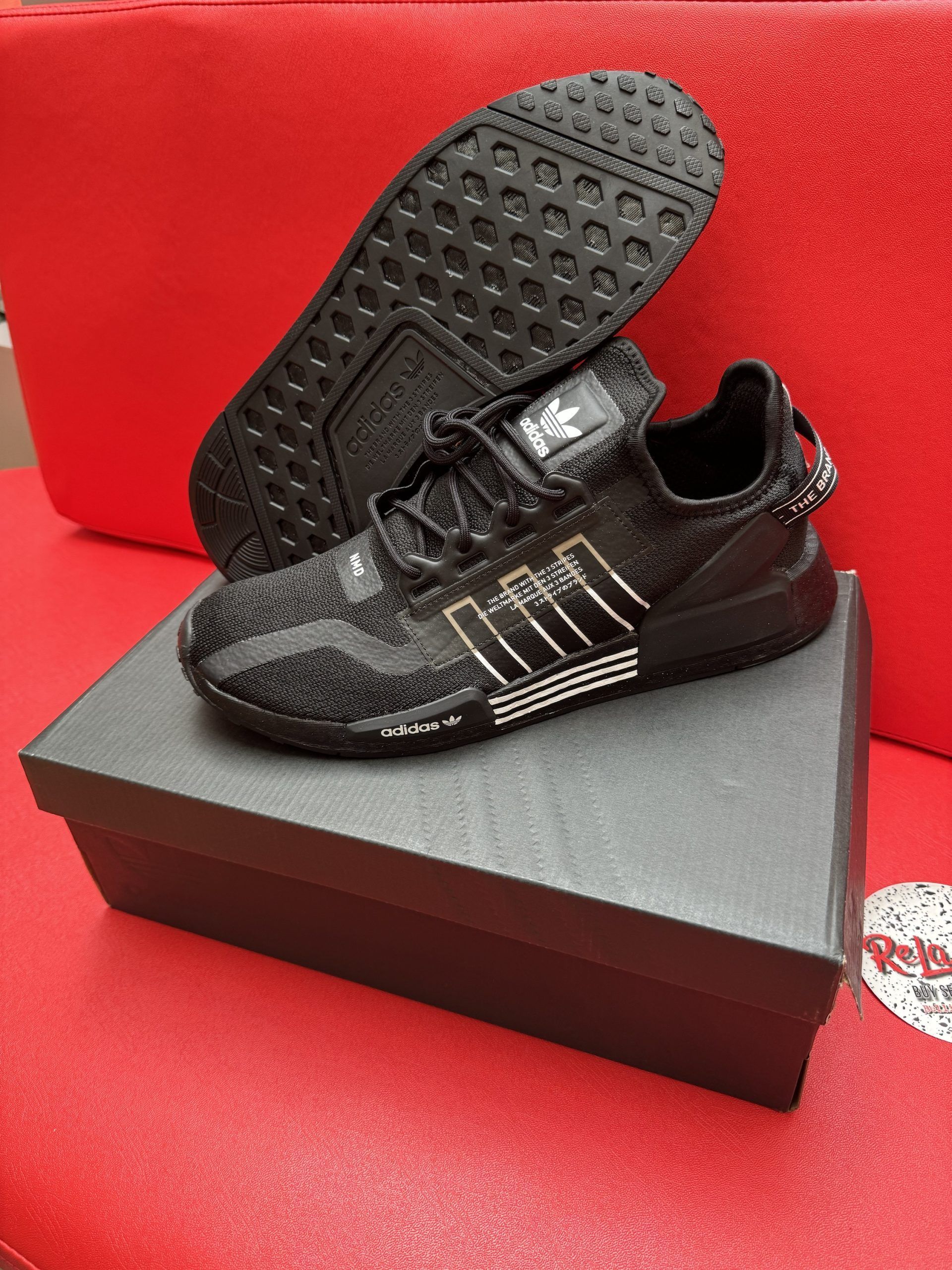 Black Adidas sneakers on a black box, with the sole visible; set on a red surface.