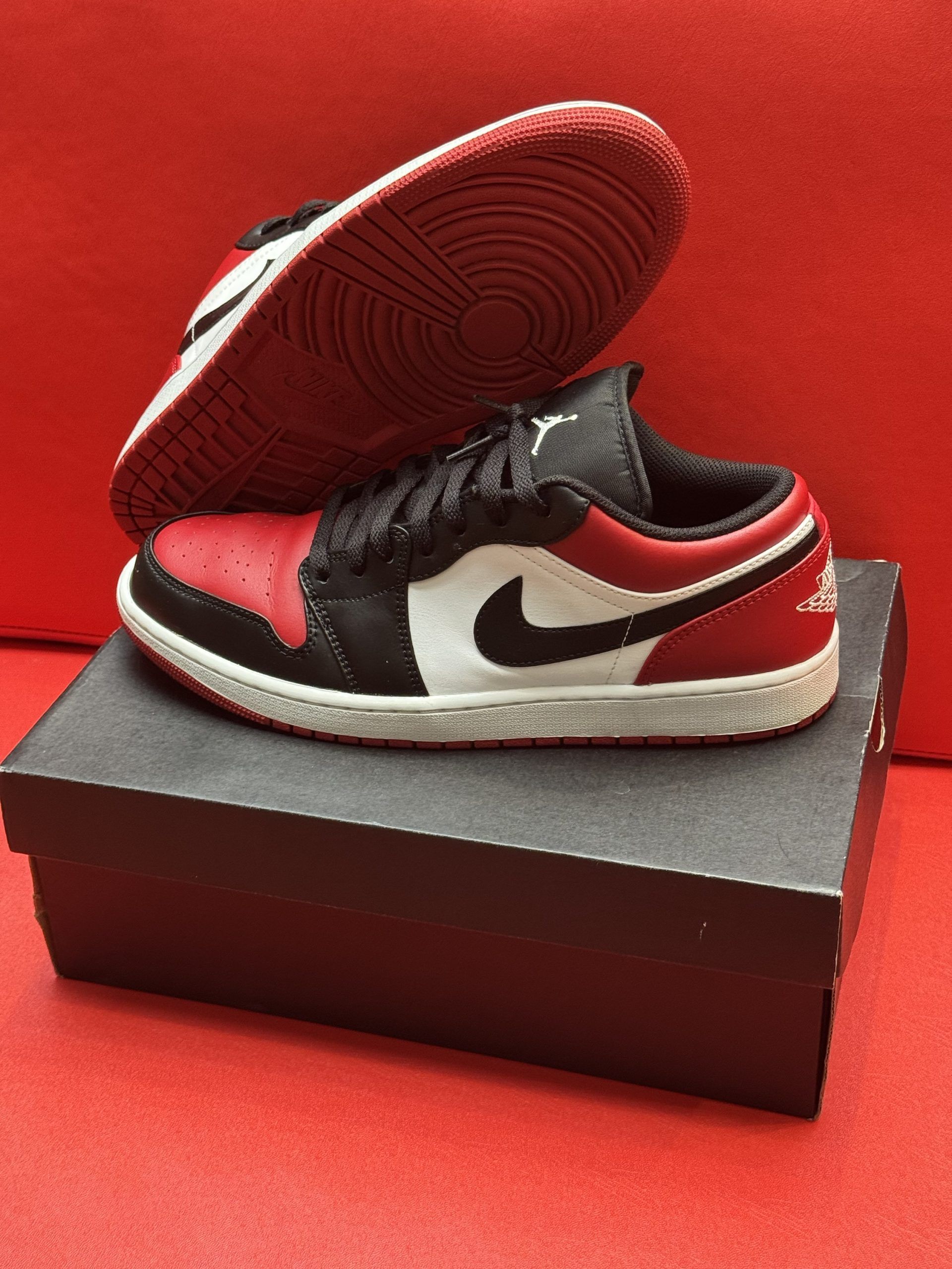 Red, black, and white Air Jordan 1 Low sneakers on a black shoebox, against a red background.