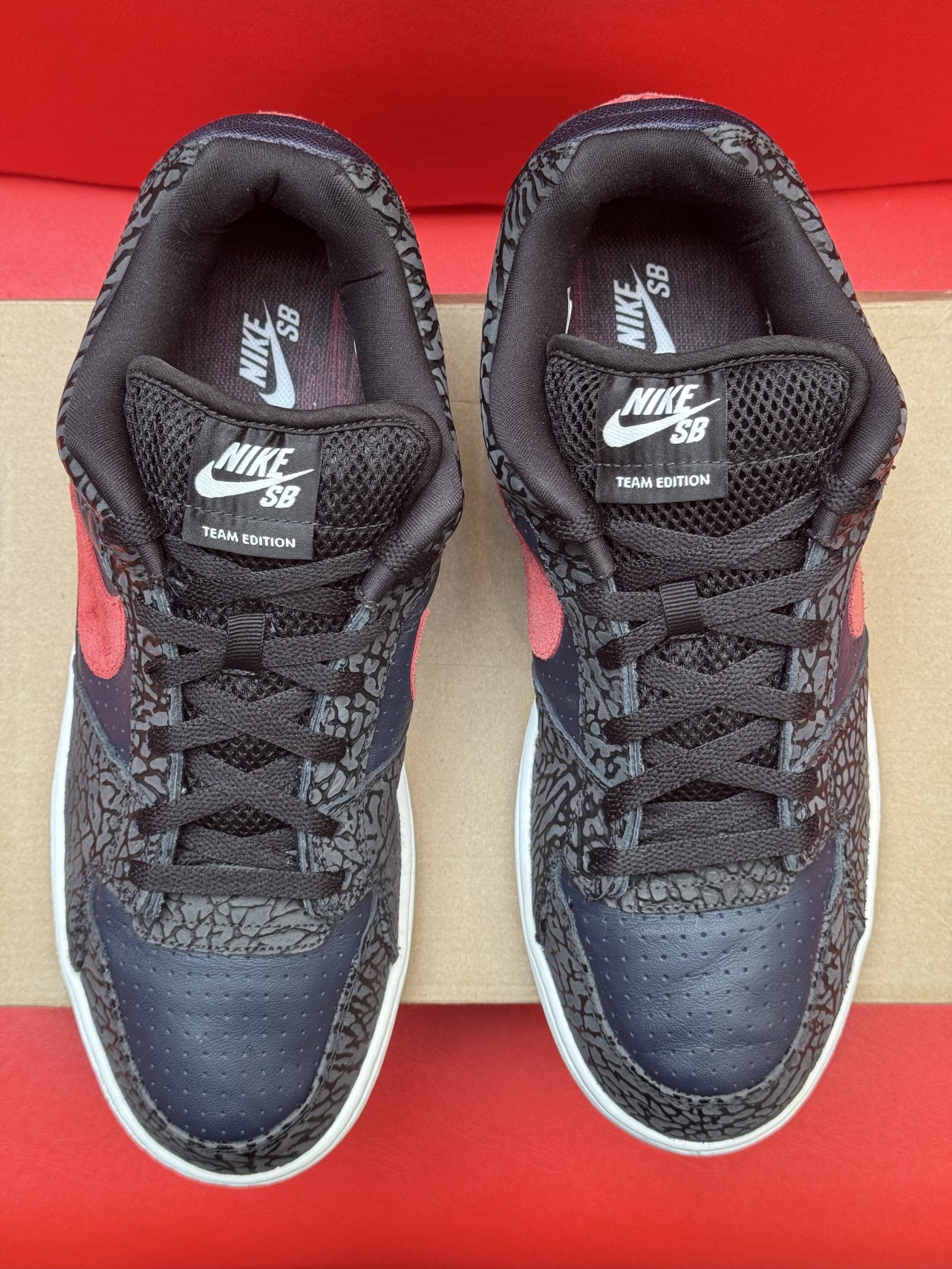 Nike Court Vision low-top sneakers in navy and red, set on a cardboard box against a red background.