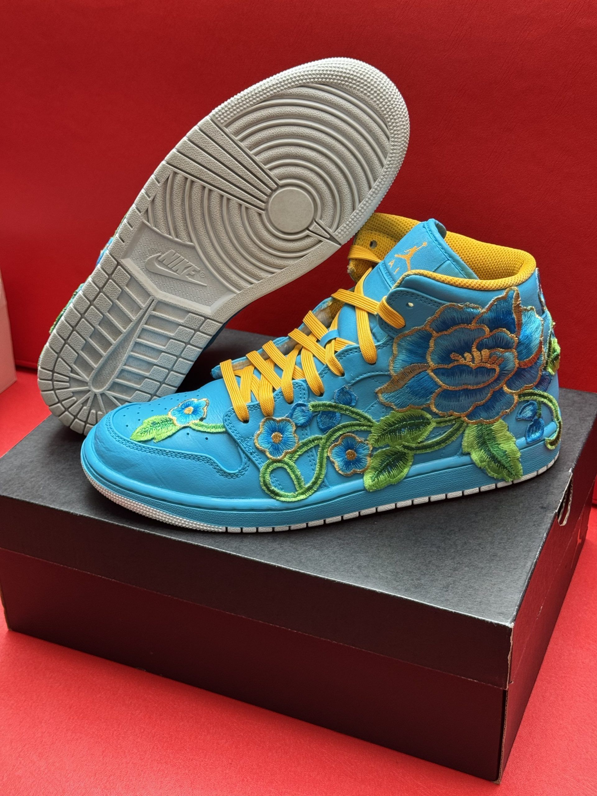 Blue and gold embroidered sneaker, floral design, on a black box, with the sole visible.