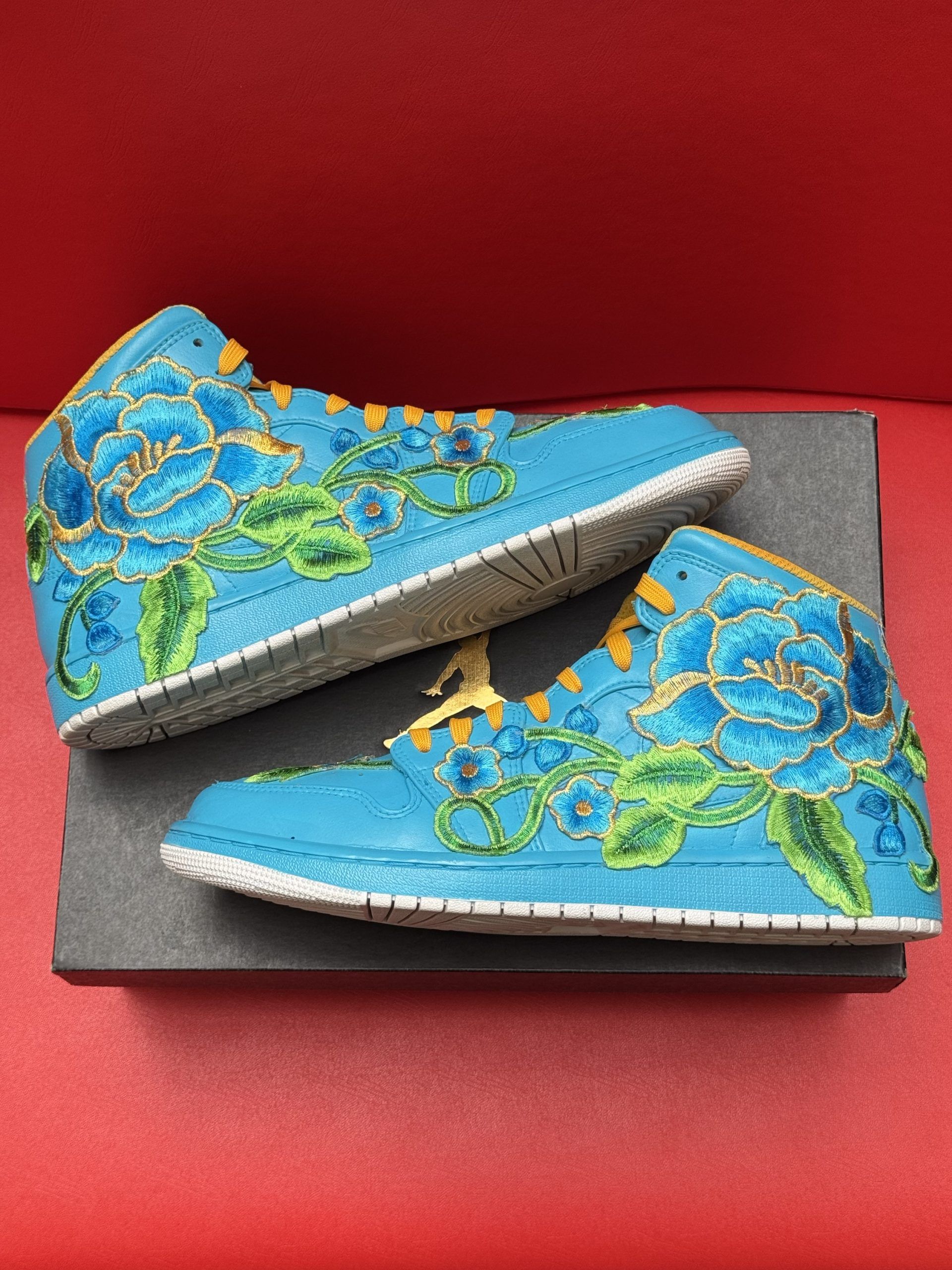Blue and gold embroidered sneaker, floral design, on a black box, with the sole visible.