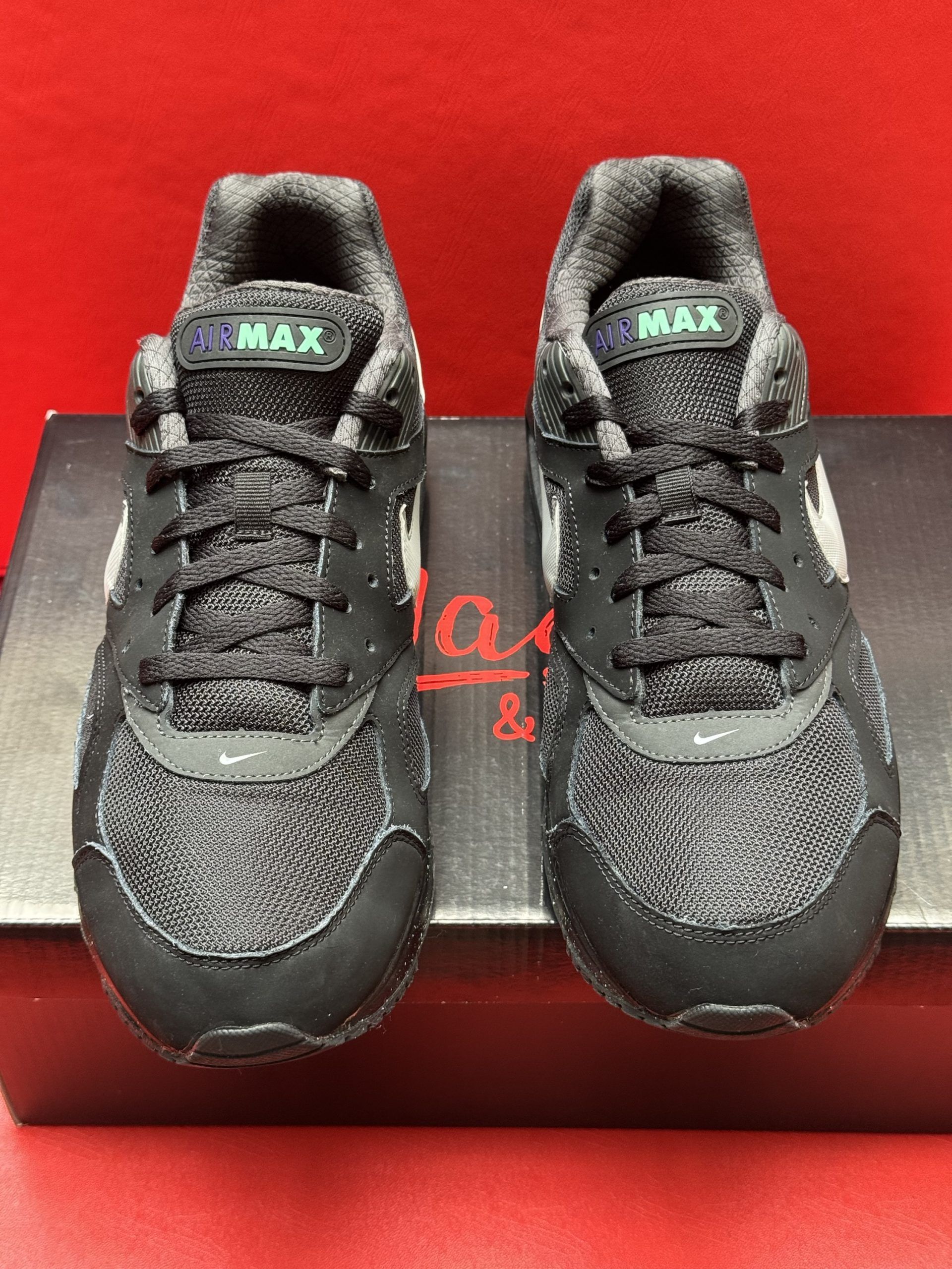 Black Nike Air Max sneakers on a black box, one shoe's sole visible, set against a red background.