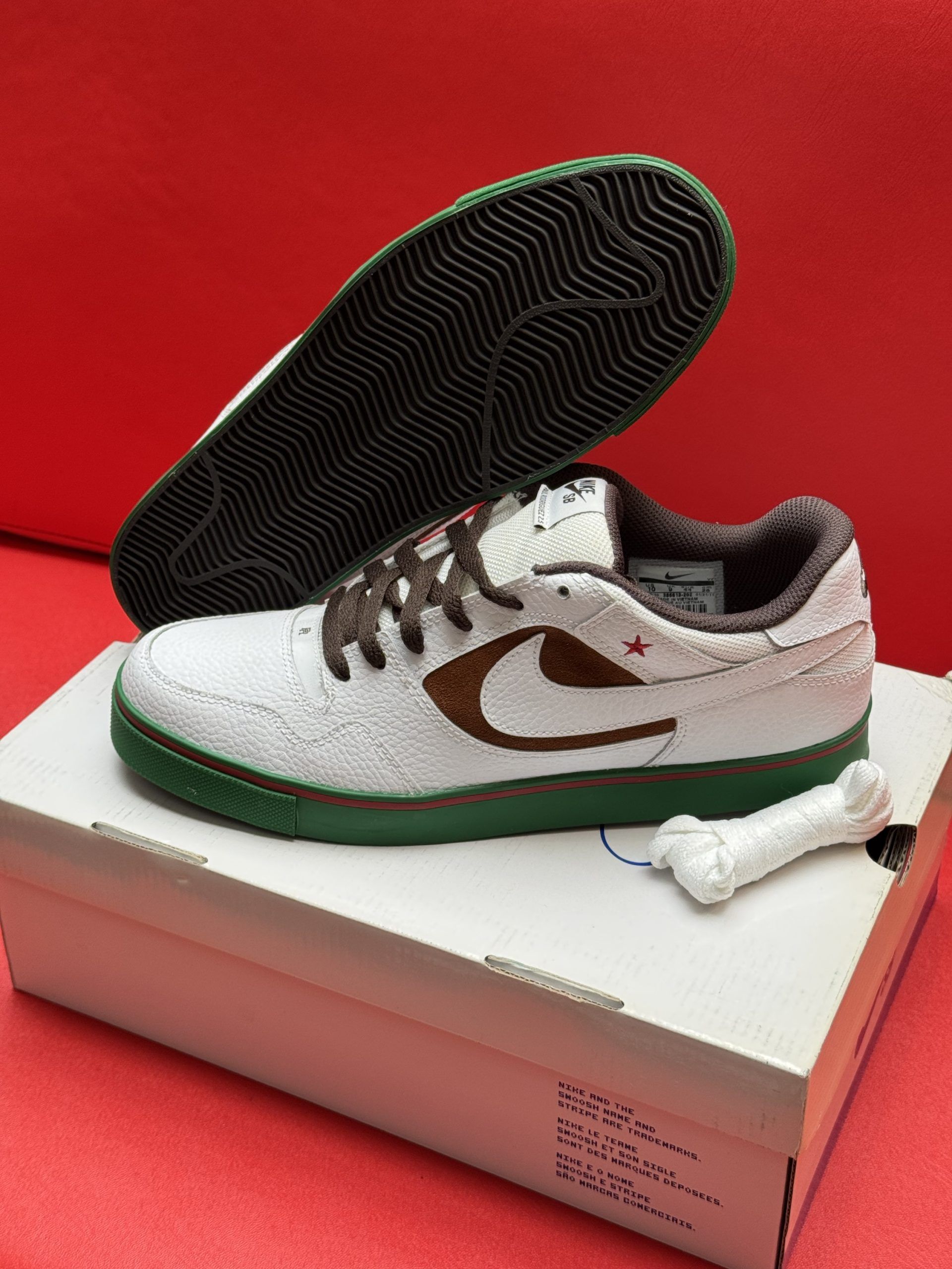 White Nike sneakers with brown swoosh and laces, green sole, bone accessory, on a box, red background.