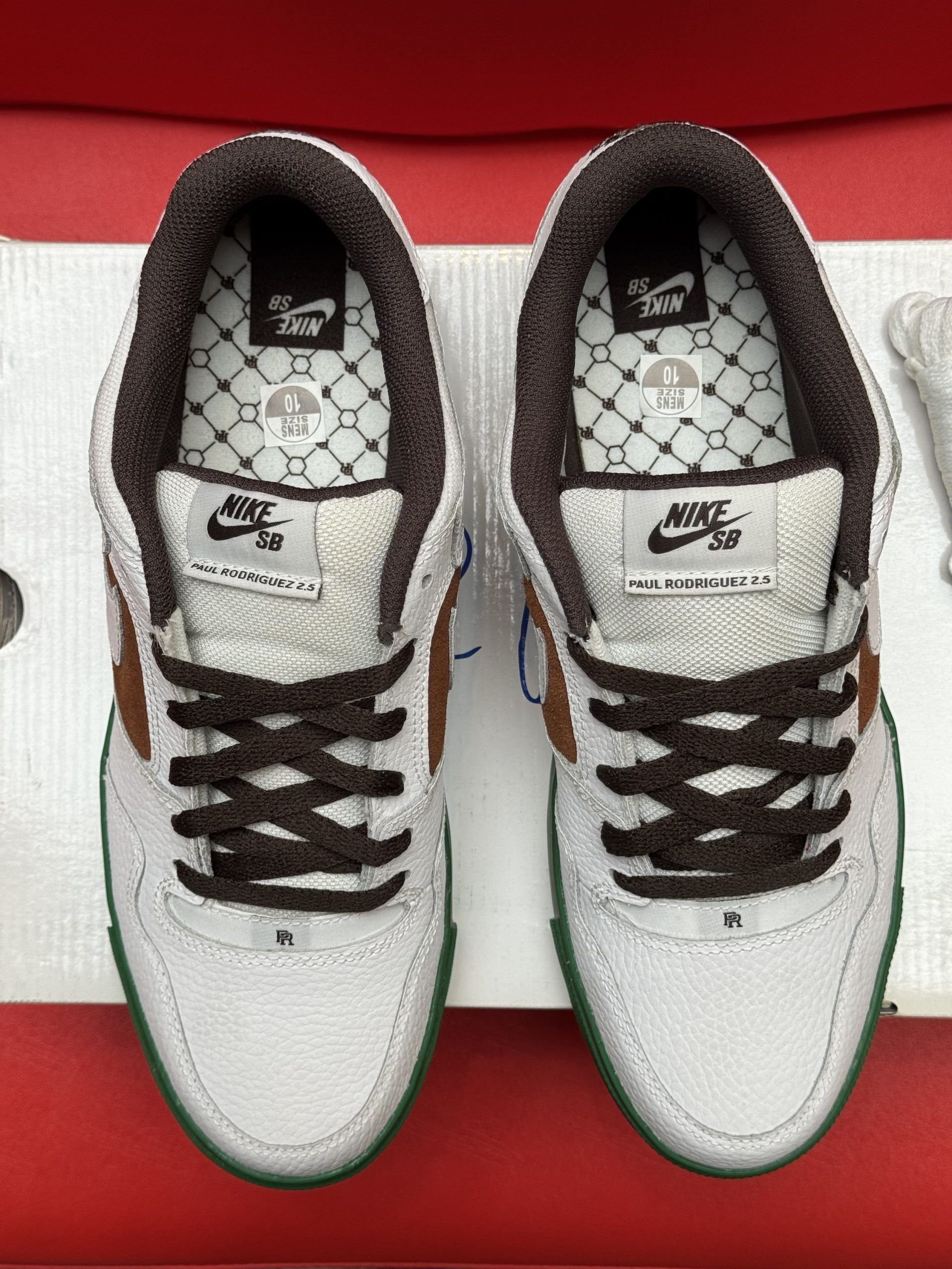White Nike sneakers with brown swoosh and laces, green sole, bone accessory, on a box, red background.