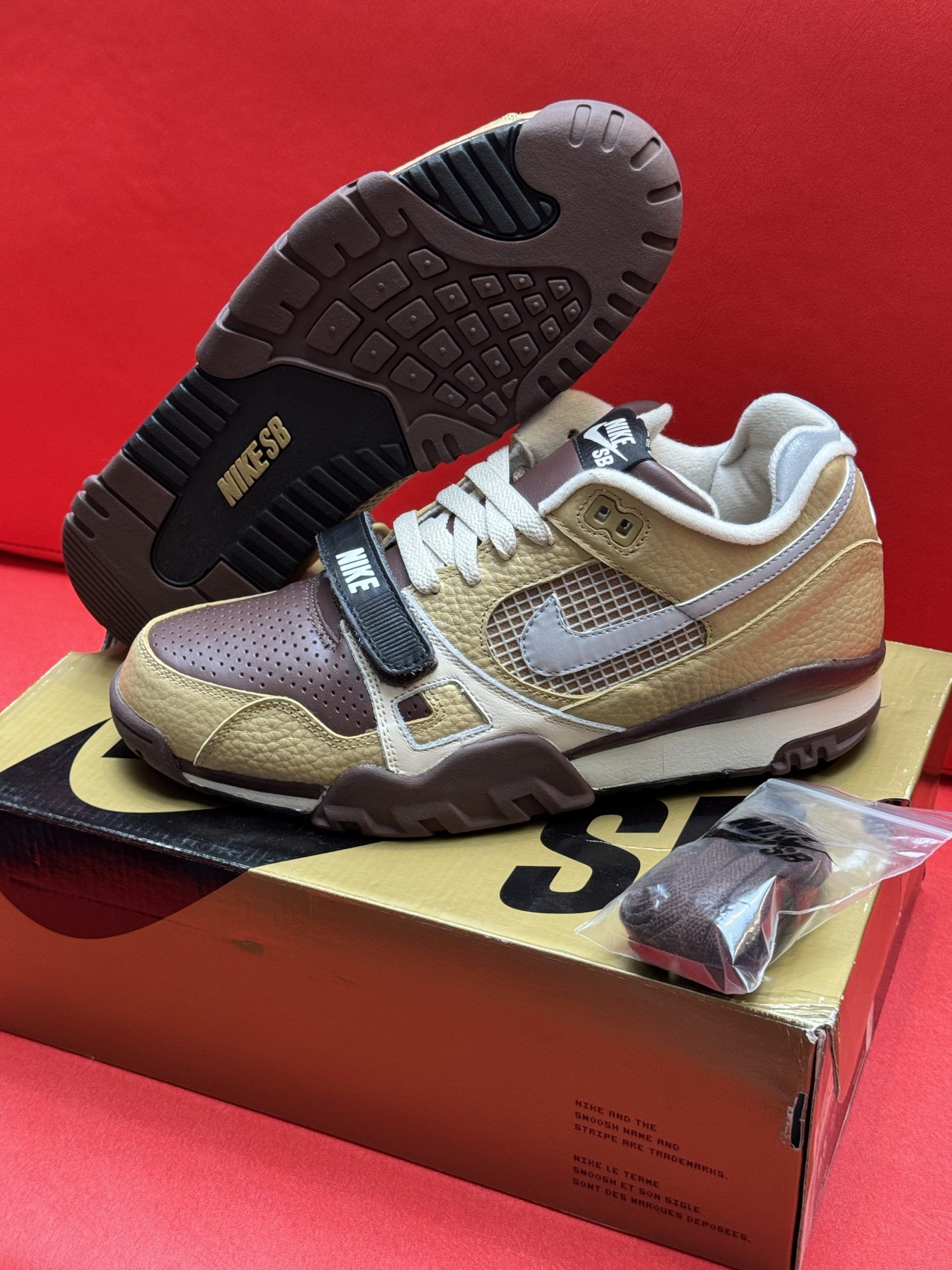 Nike Air Trainer 1 sneakers in brown and tan, on a box with an extra pair of laces. The sole is visible.