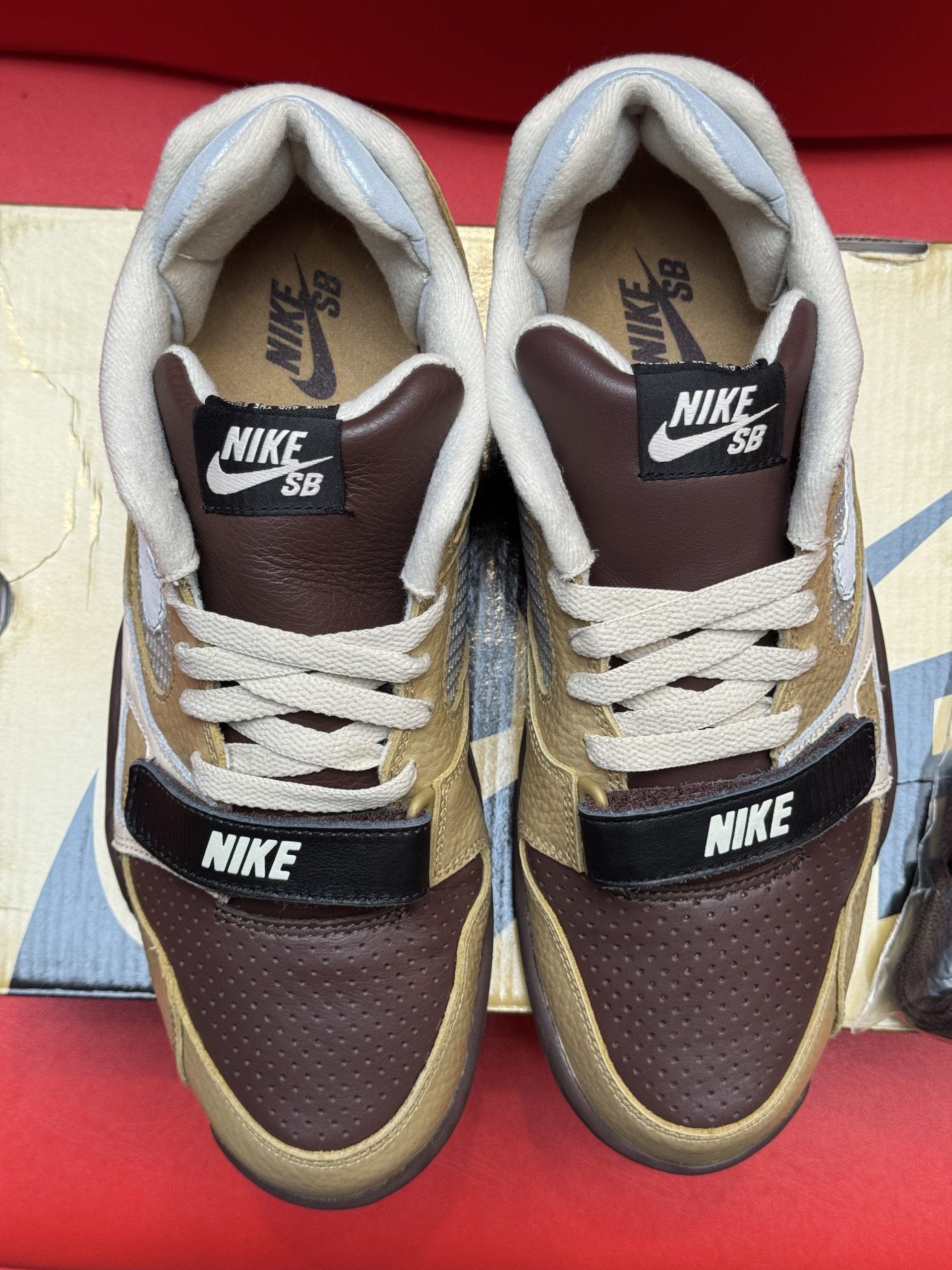 Nike Air Trainer 1 sneakers in brown and tan, on a box with an extra pair of laces. The sole is visible.