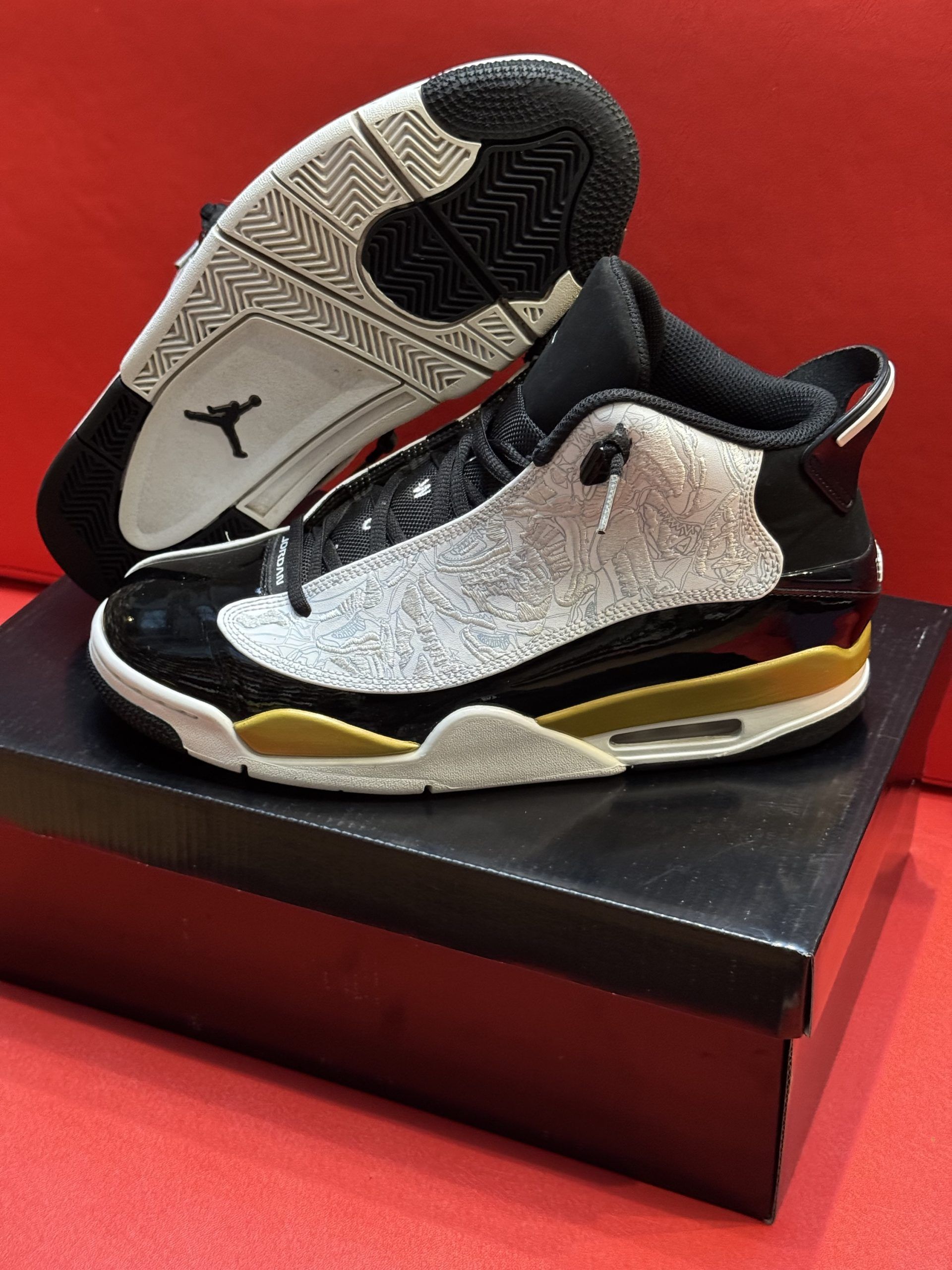 Black and white Air Jordan sneakers with gold accents, displayed on a black box against a red background.