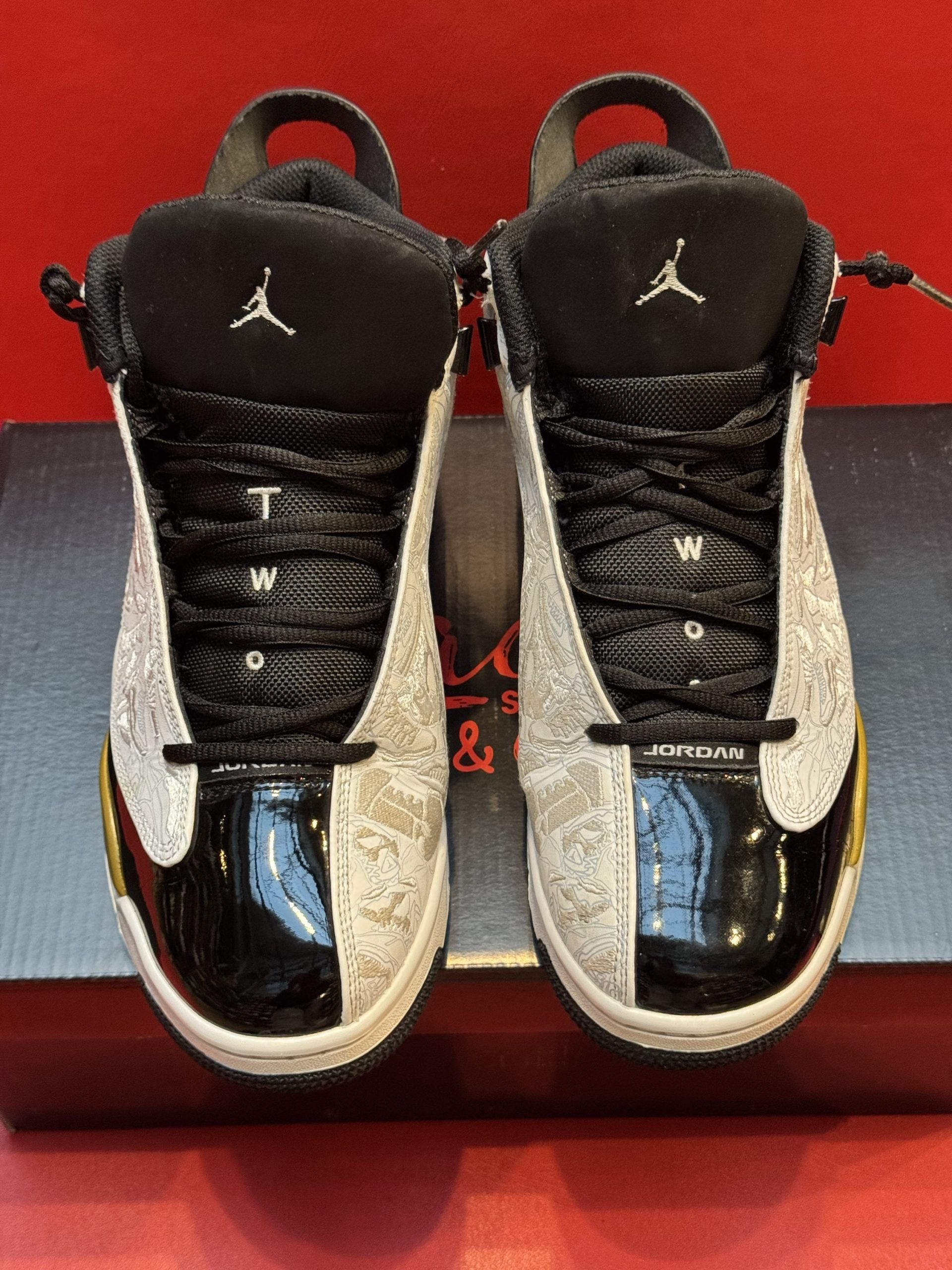 Black and white Air Jordan sneakers with gold accents, displayed on a black box against a red background.