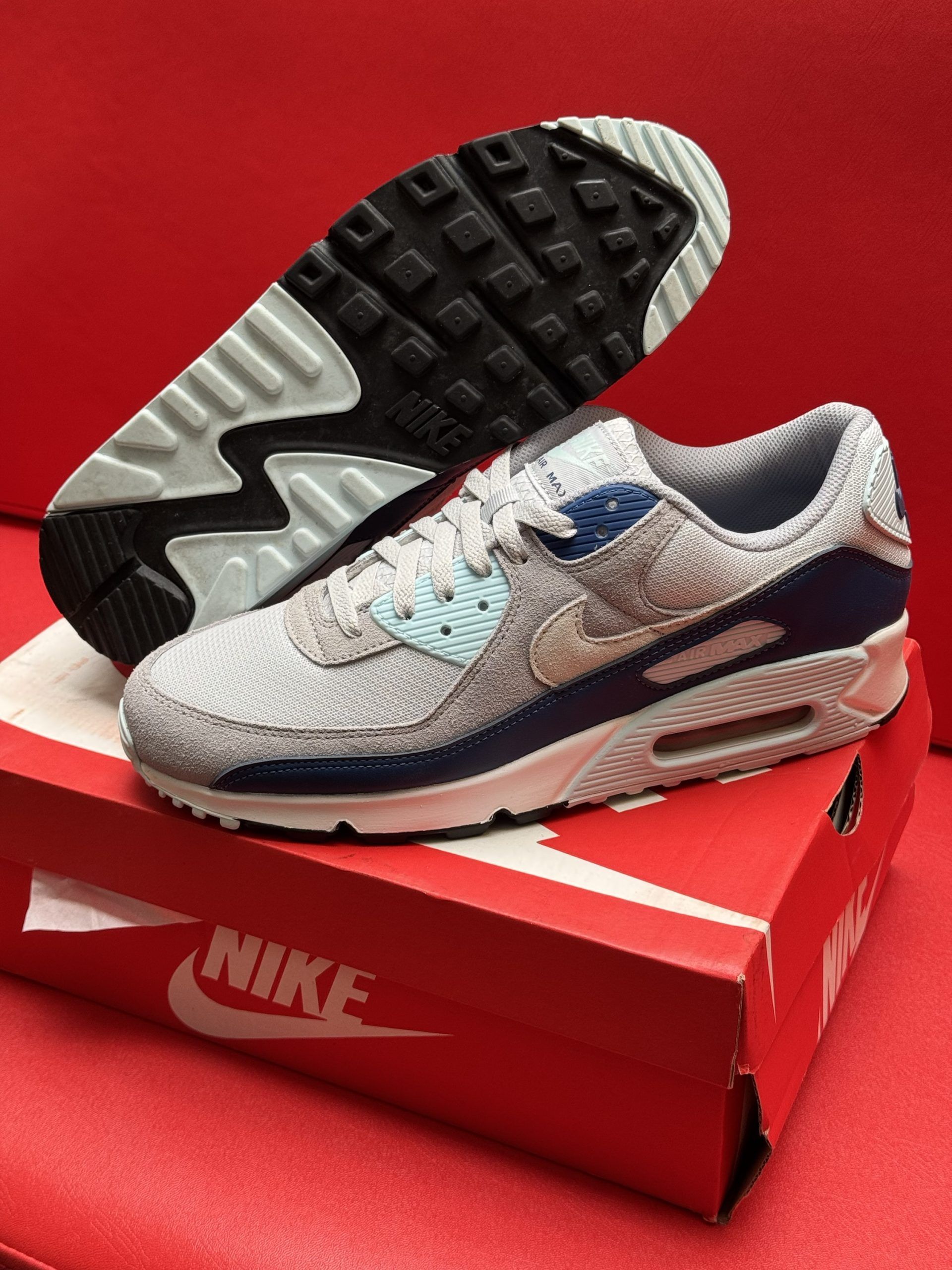 Nike Air Max 90 sneakers in gray, blue, and beige, with textured upper, atop a red Nike box and sole displayed.