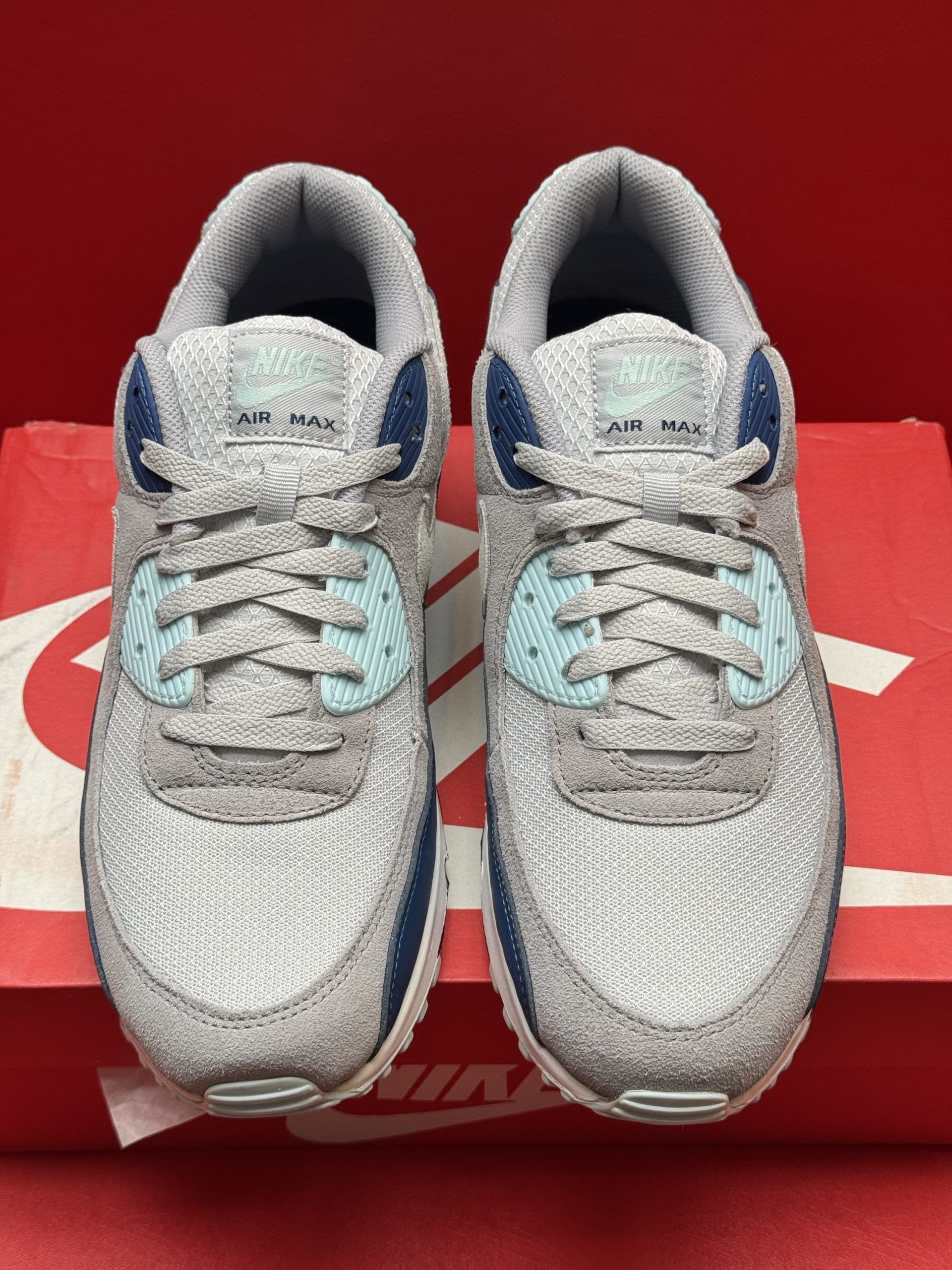 Nike Air Max 90 sneakers in gray, blue, and beige, with textured upper, atop a red Nike box and sole displayed.