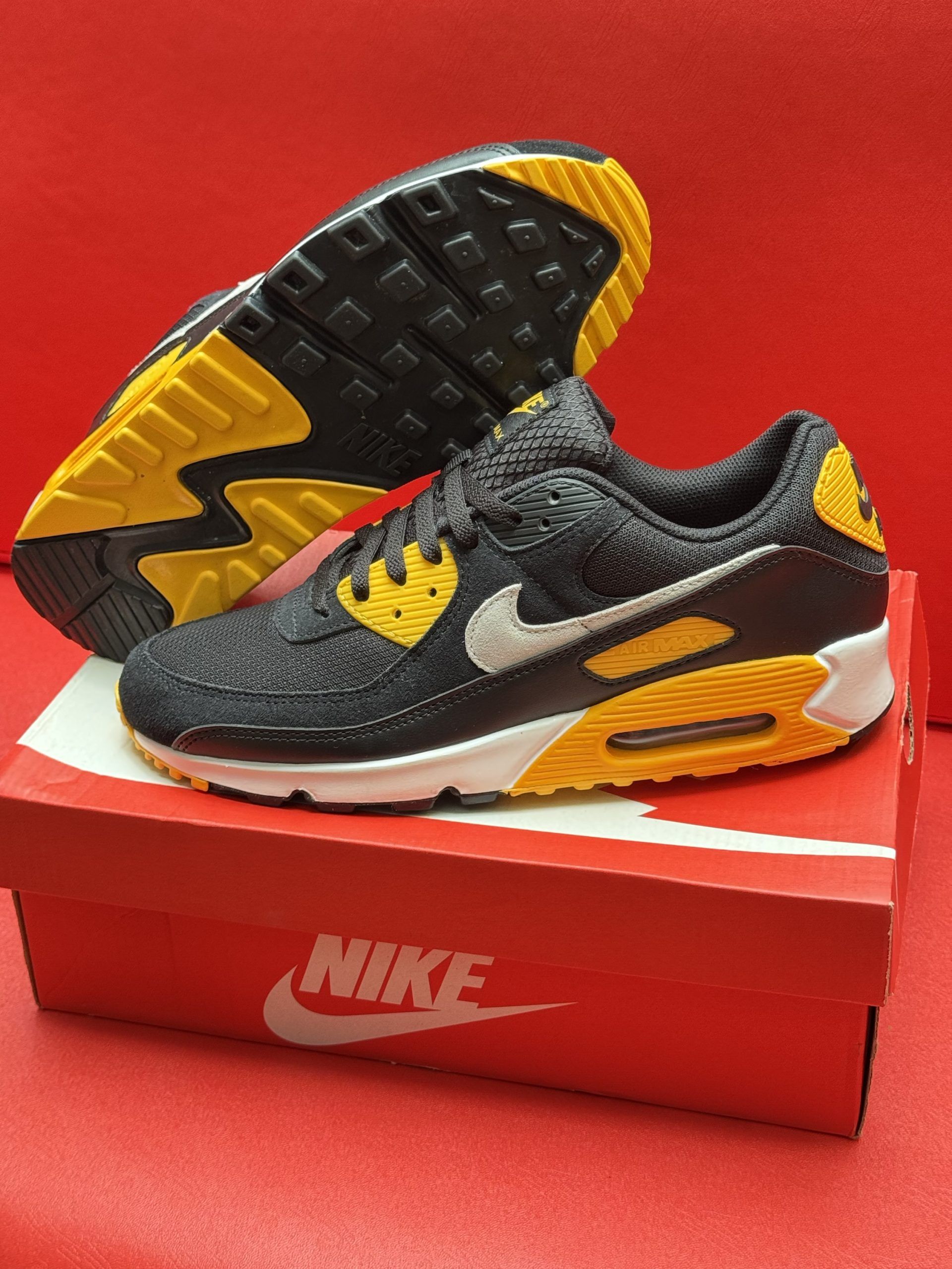 Black and yellow Nike Air Max 90 sneakers on a red Nike box, with the sole visible.