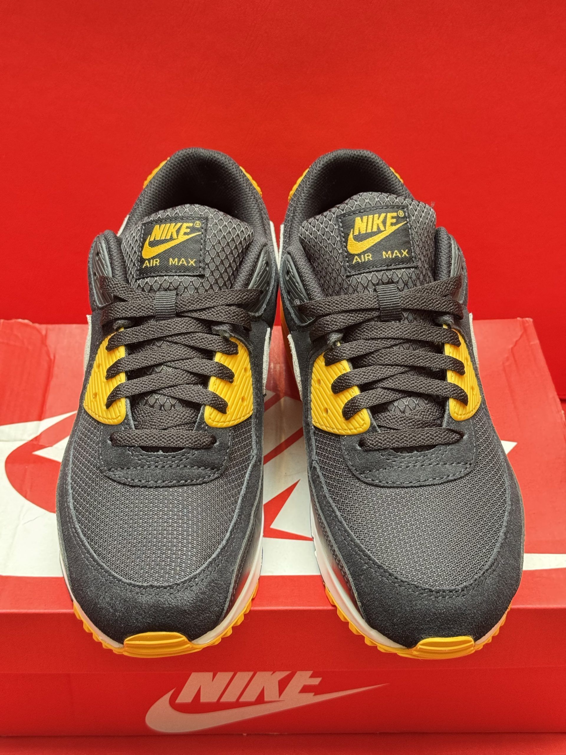 Black and yellow Nike Air Max 90 sneakers on a red Nike box, with the sole visible.