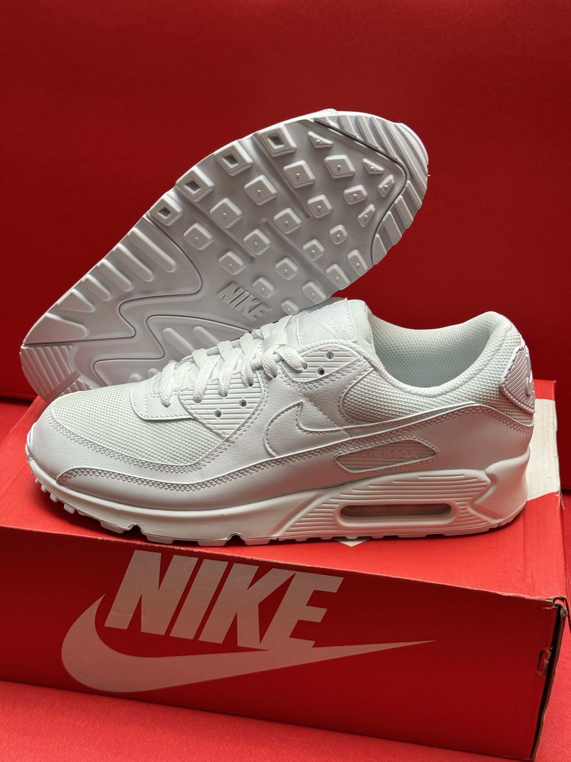 White Nike Air Max 90 sneakers on a red Nike box, with the sole visible.