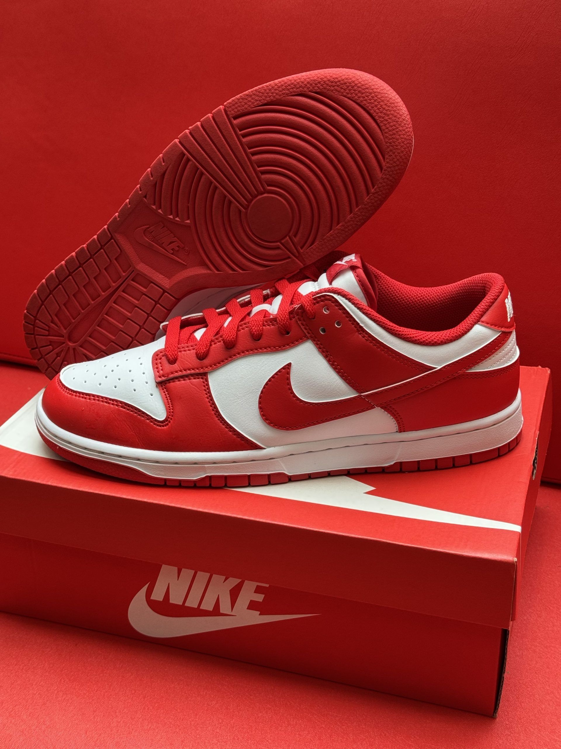 Red and white Nike Dunk Low sneakers with the sole visible, on a red Nike box, against a red background.