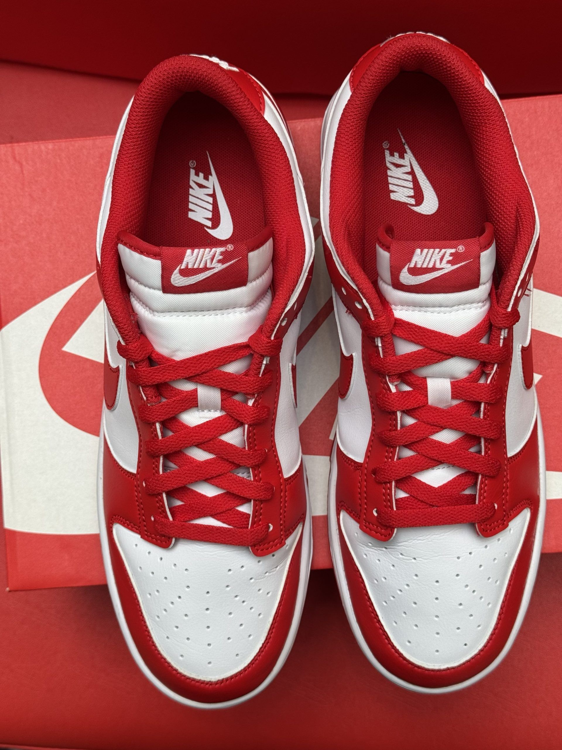 Red and white Nike Dunk Low sneakers with the sole visible, on a red Nike box, against a red background.