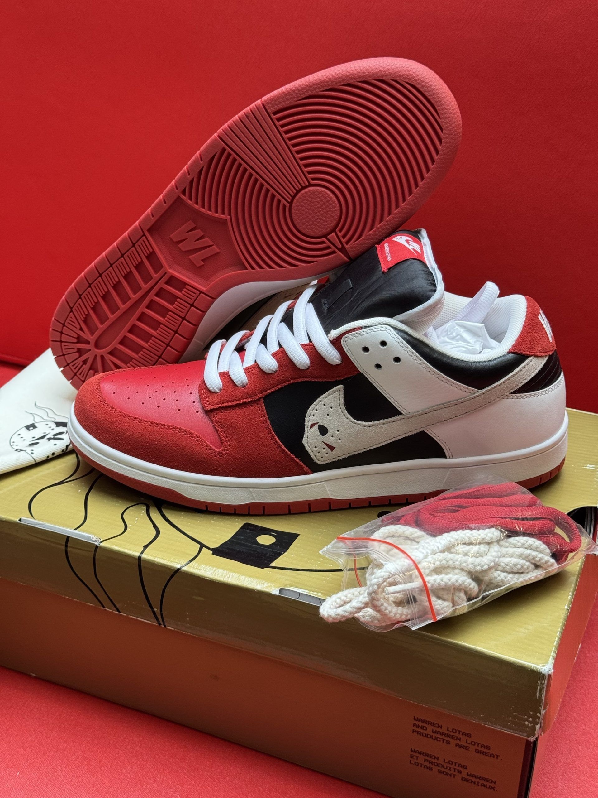 Red, white, and black Nike Dunk Low sneakers with extra white laces, sitting on a shoebox against a red backdrop.