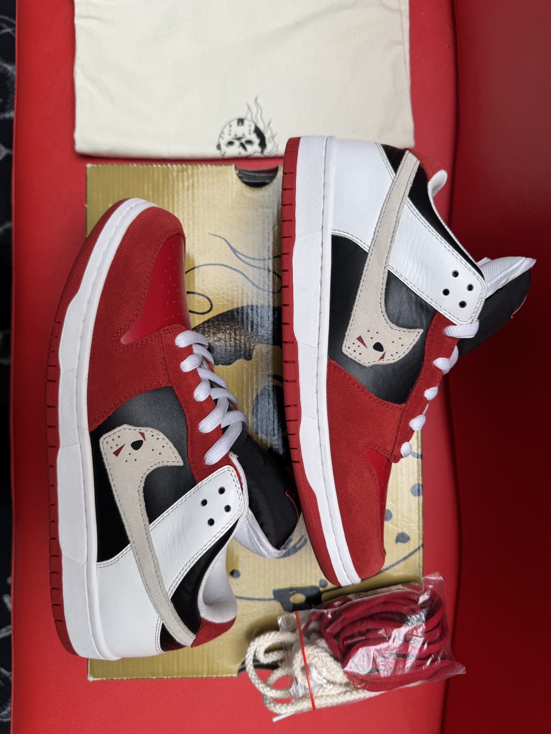 Red, white, and black Nike Dunk Low sneakers with extra white laces, sitting on a shoebox against a red backdrop.