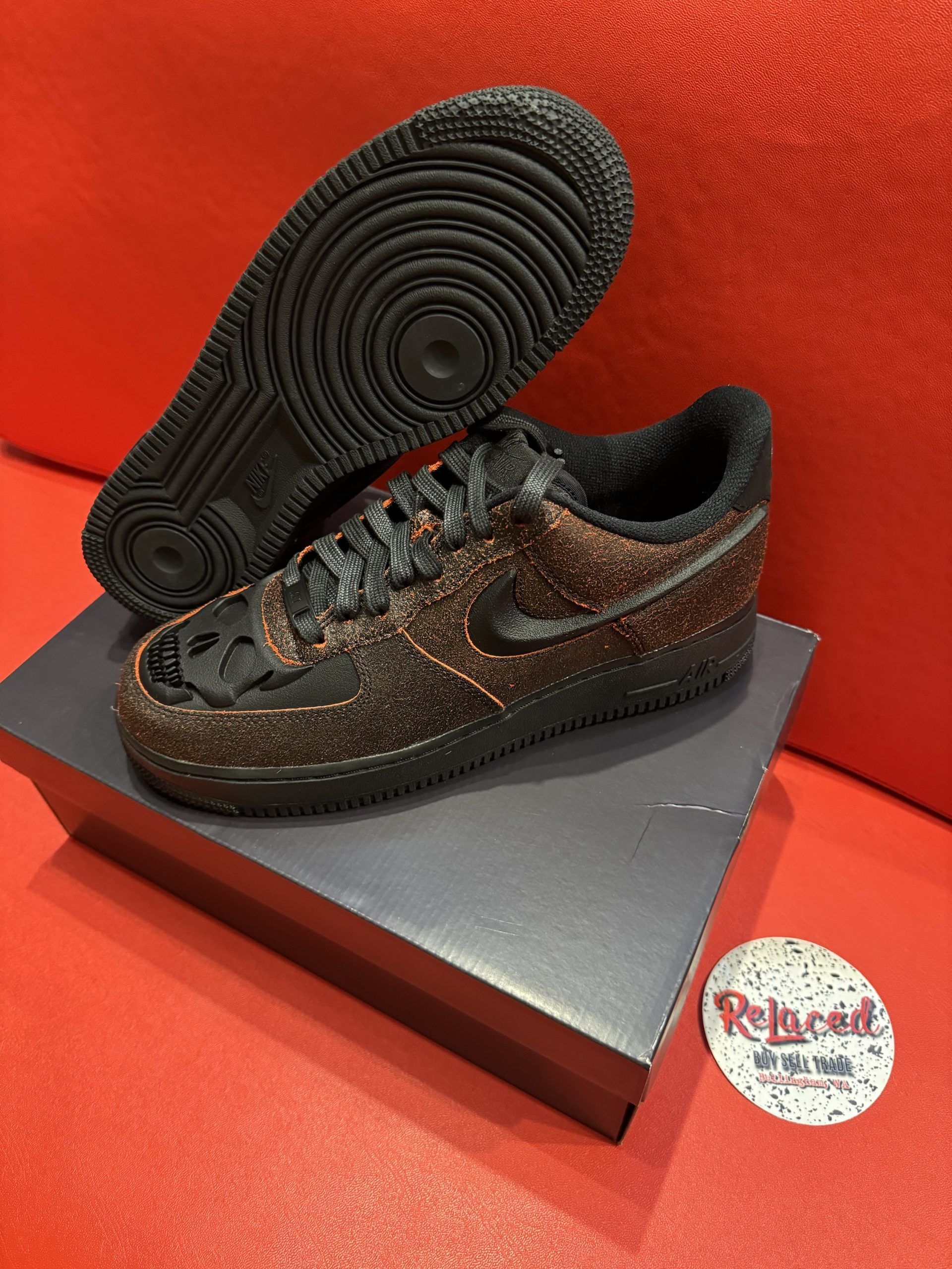 Black and bronze Nike Air Force 1 sneakers with textured soles, displayed on a black box against a red surface.