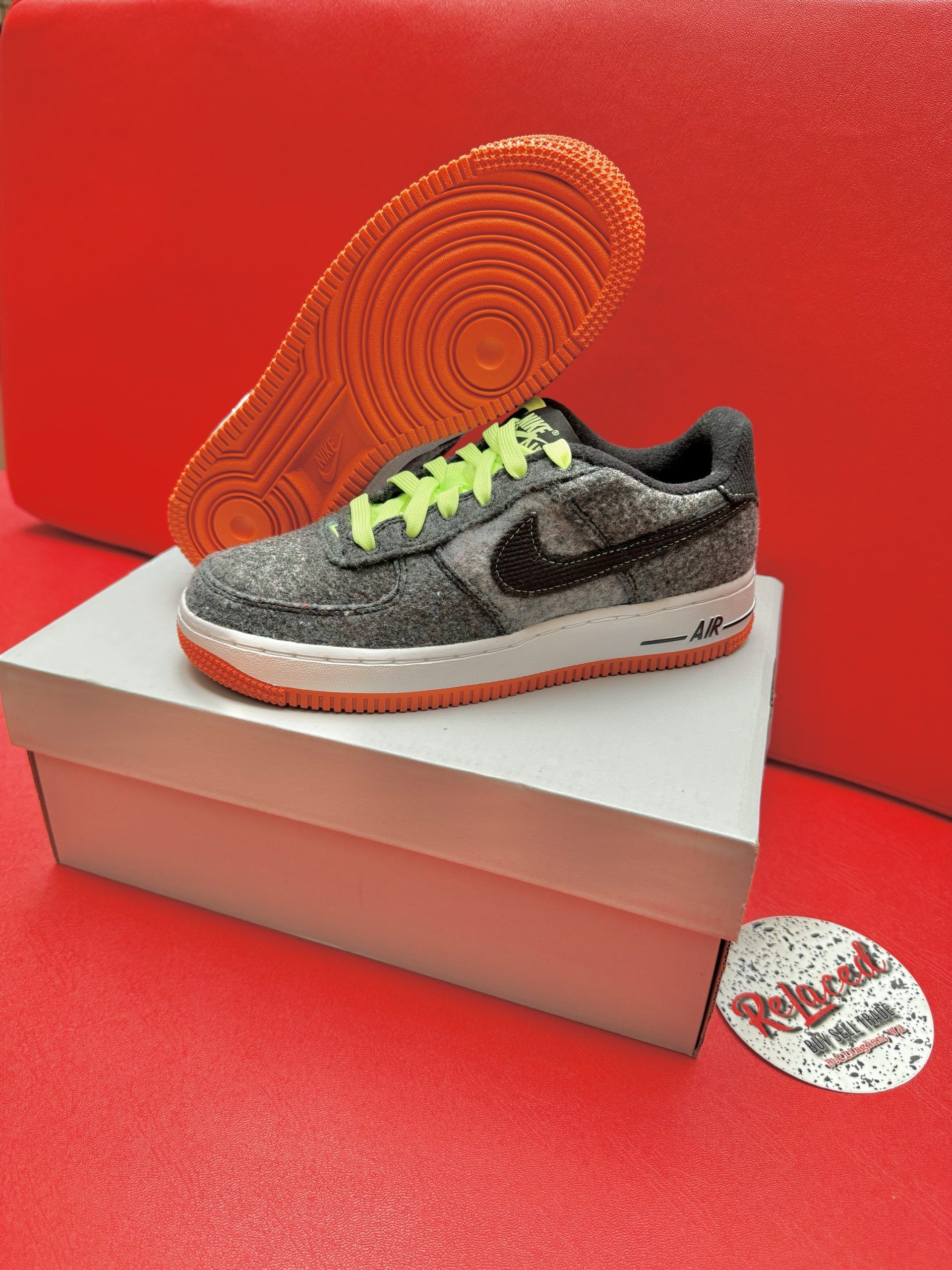 Gray Nike Air Force 1 sneaker with neon laces and orange sole, resting on a white box on a red surface.