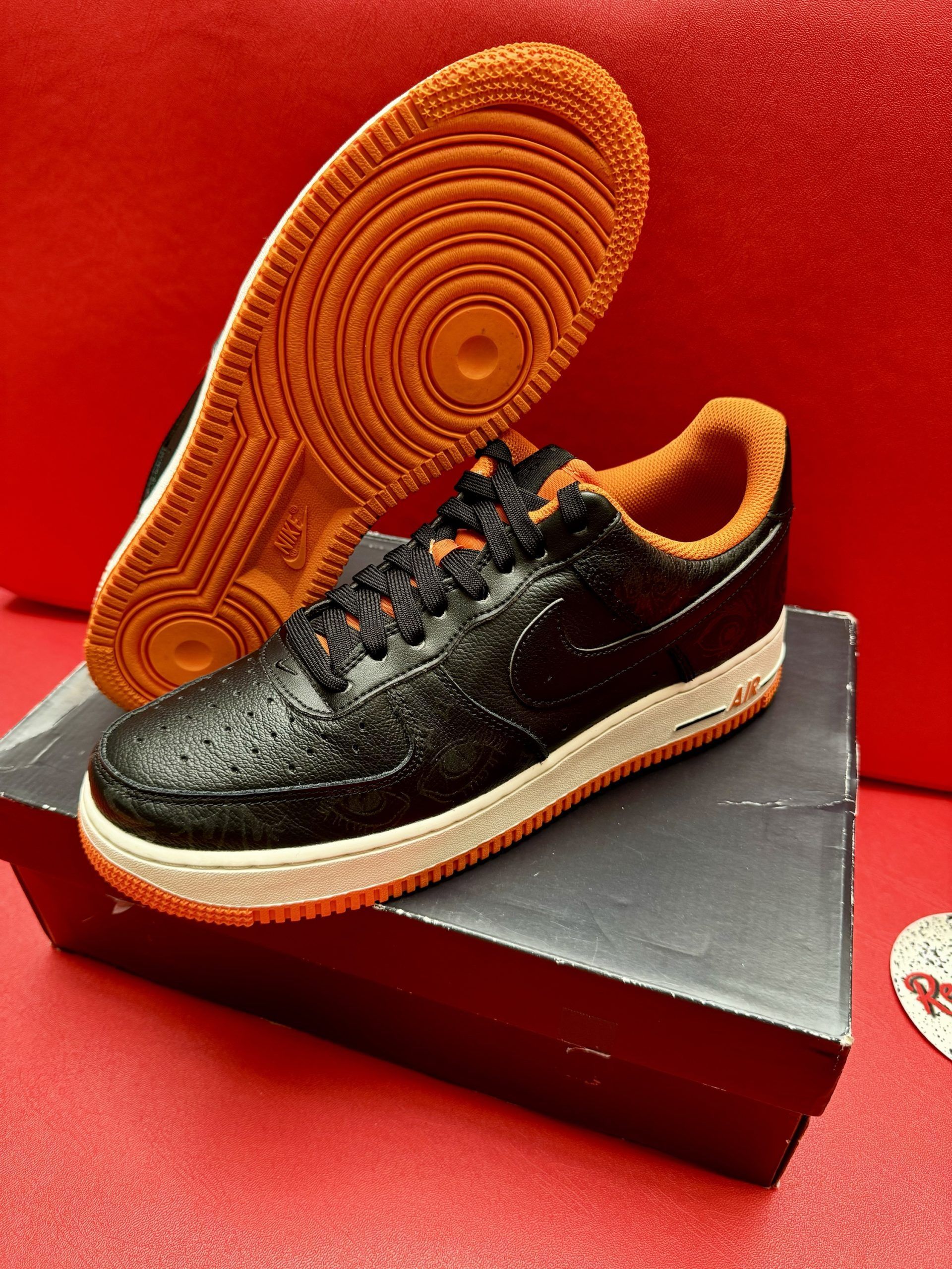 Black Nike Air Force 1 sneakers with orange accents, on a box against a red backdrop.