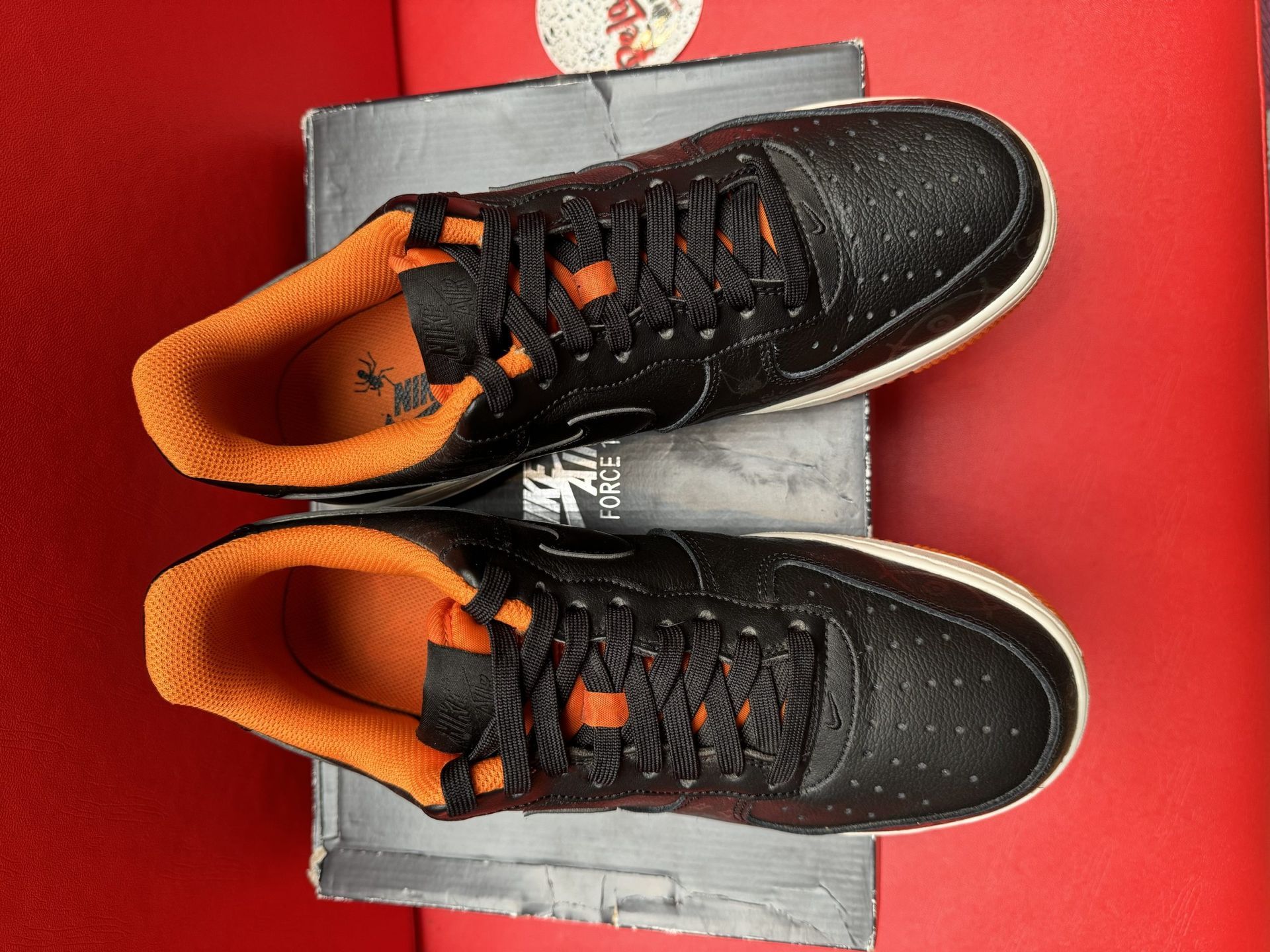 Black Nike Air Force 1 sneakers with orange accents, on a box against a red backdrop.
